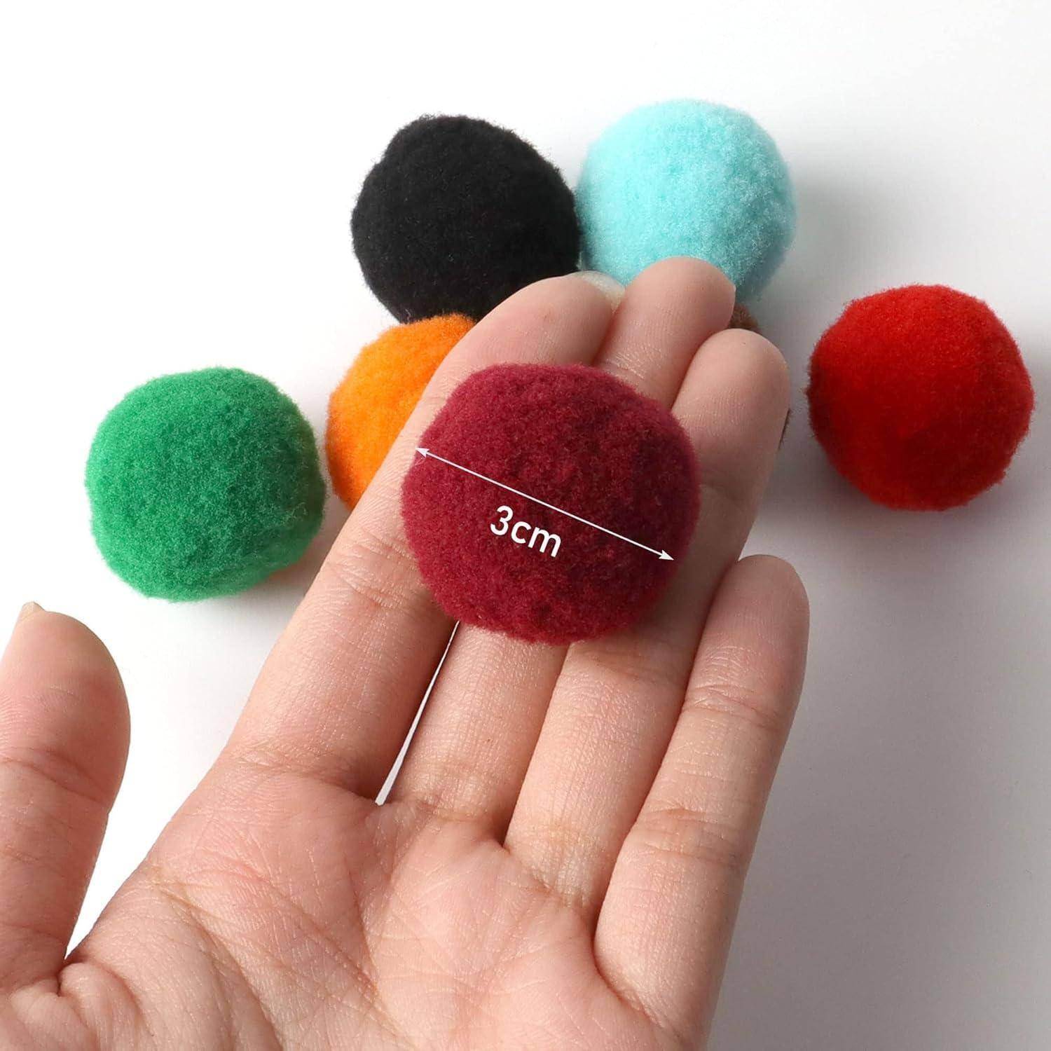 Eine Hand hält einen roten Pompom, der mit '3 cm' beschriftet ist. Fünf mehrfarbige Pompoms liegen im Hintergrund auf einer weißen Oberfläche.