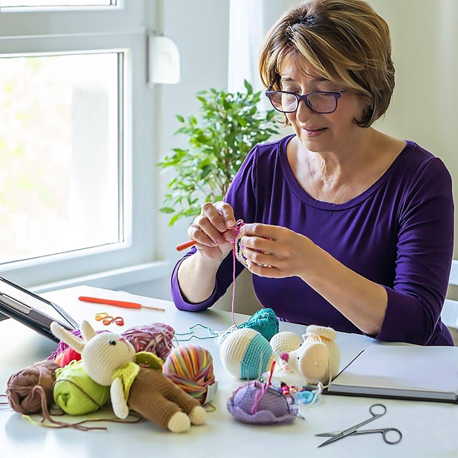 Eine Frau in einem lila Shirt sitzt an einem Tisch und häkelt bunte Stofftiere. Scheren und Garn liegen in der Nähe. Im Hintergrund befindet sich ein Fenster und eine Pflanze.