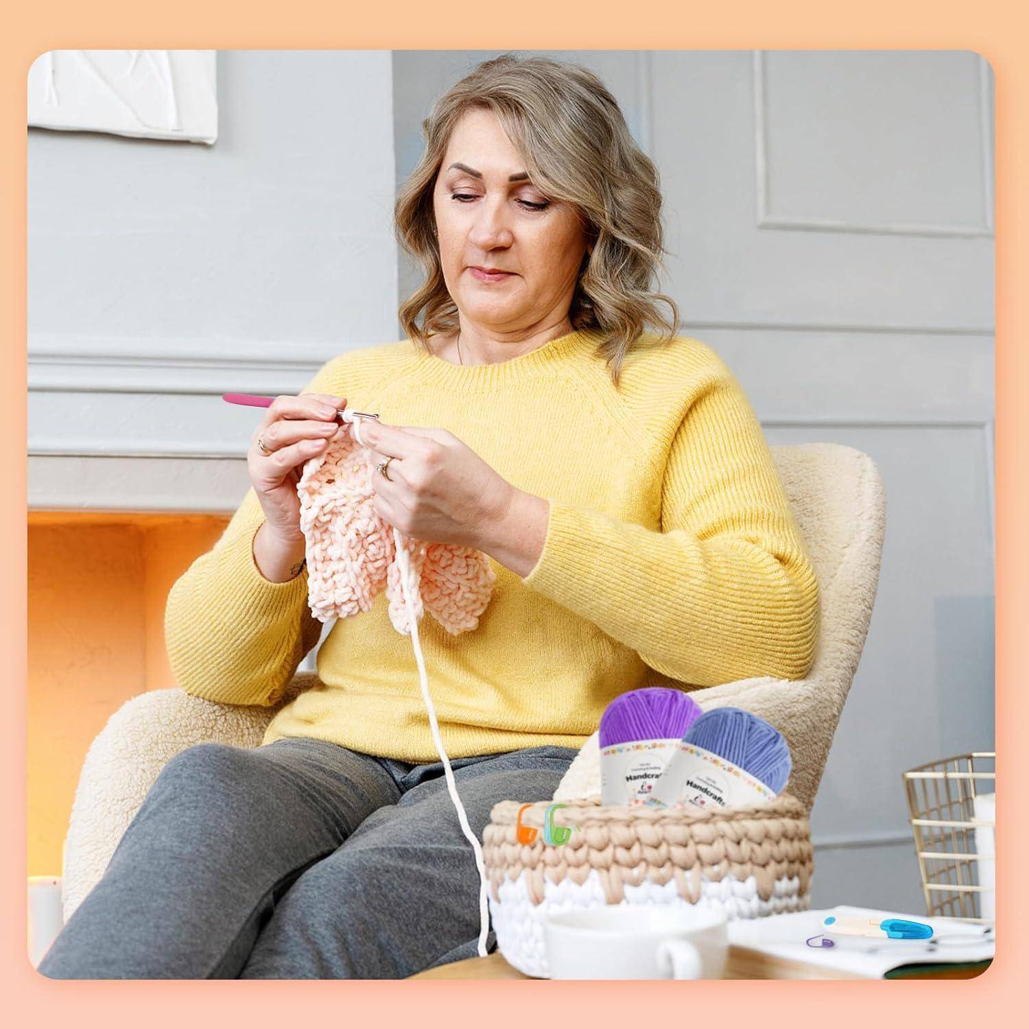 Eine Person sitzt auf einem Stuhl und strickt mit rosa Garn. Ein Korb mit Strickutensilien steht im Vordergrund auf einem Tisch.