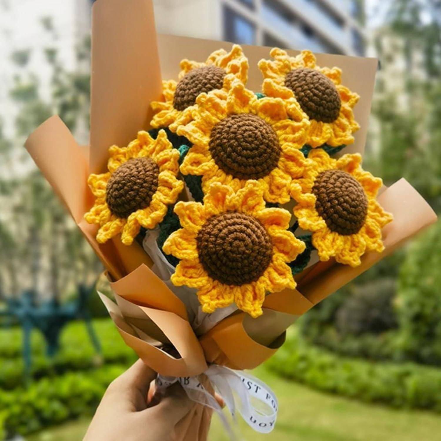 Ein Strauß gehäkelter Sonnenblumen, gehalten vor einer Parklandschaft mit Gebäuden im Hintergrund, der kunstfertiges Handwerksgeschick zur Schau stellt.