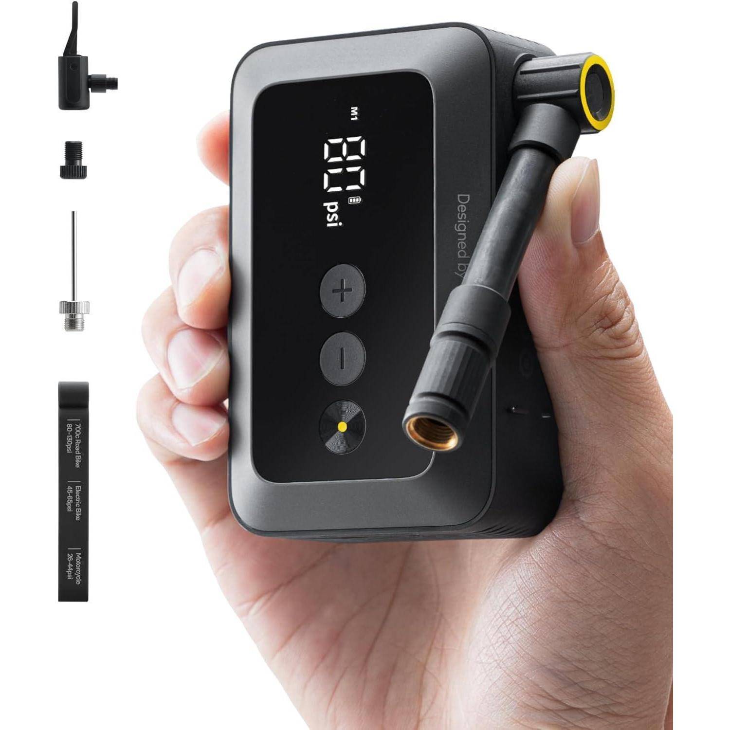 Eine Person hält einen digitalen Reifenfüller, auf dessen Display '20 psi' angezeigt wird. Verschiedene Düsenadapter sind seitlich zu sehen.