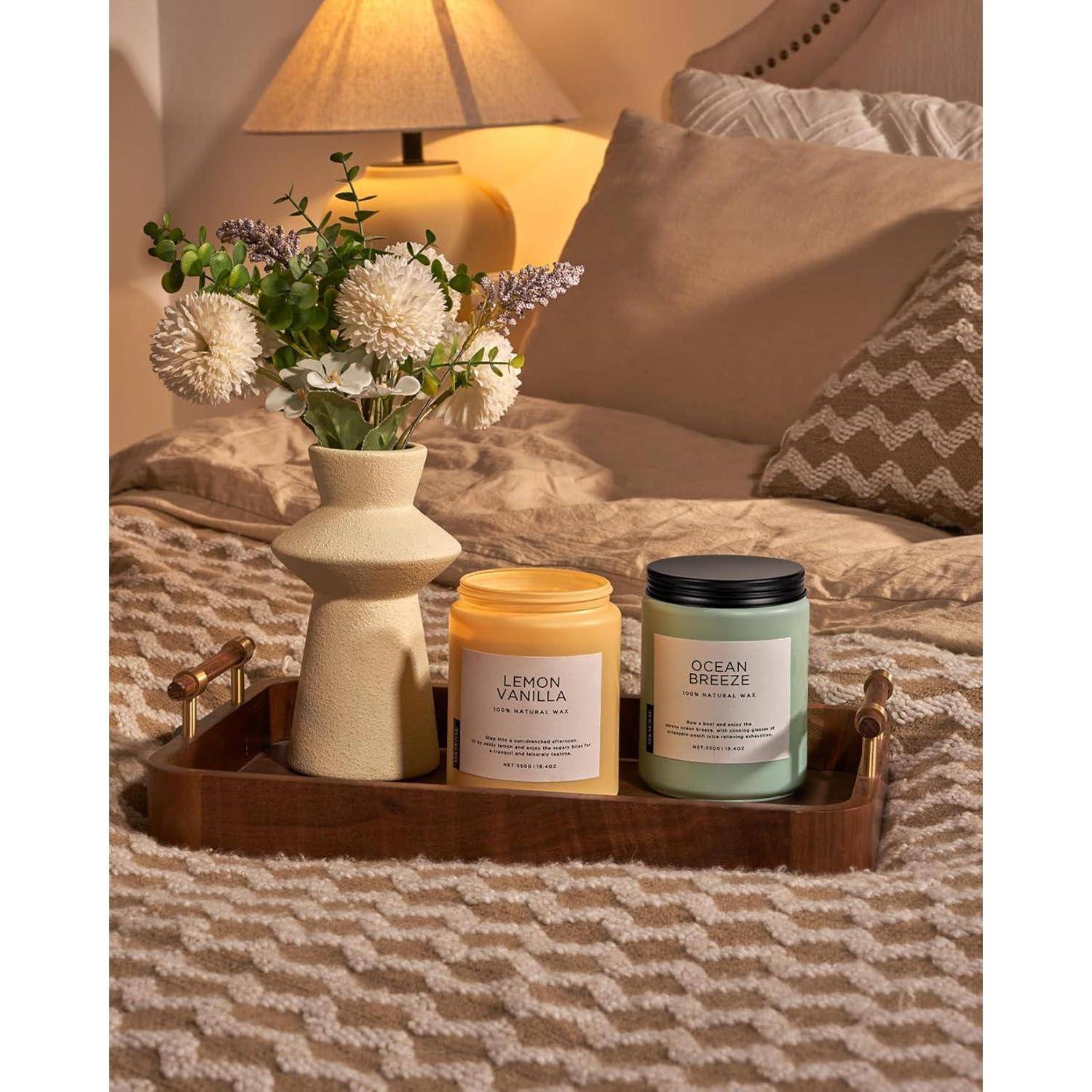 Eine gemütliche Schlafzimmerszene mit einem Holztablett auf dem Bett, das eine Vase mit weißen Blumen und zwei duftenden Kerzen mit den Bezeichnungen ‚Zitrone Vanille' und ‚Meeresluft' trägt.