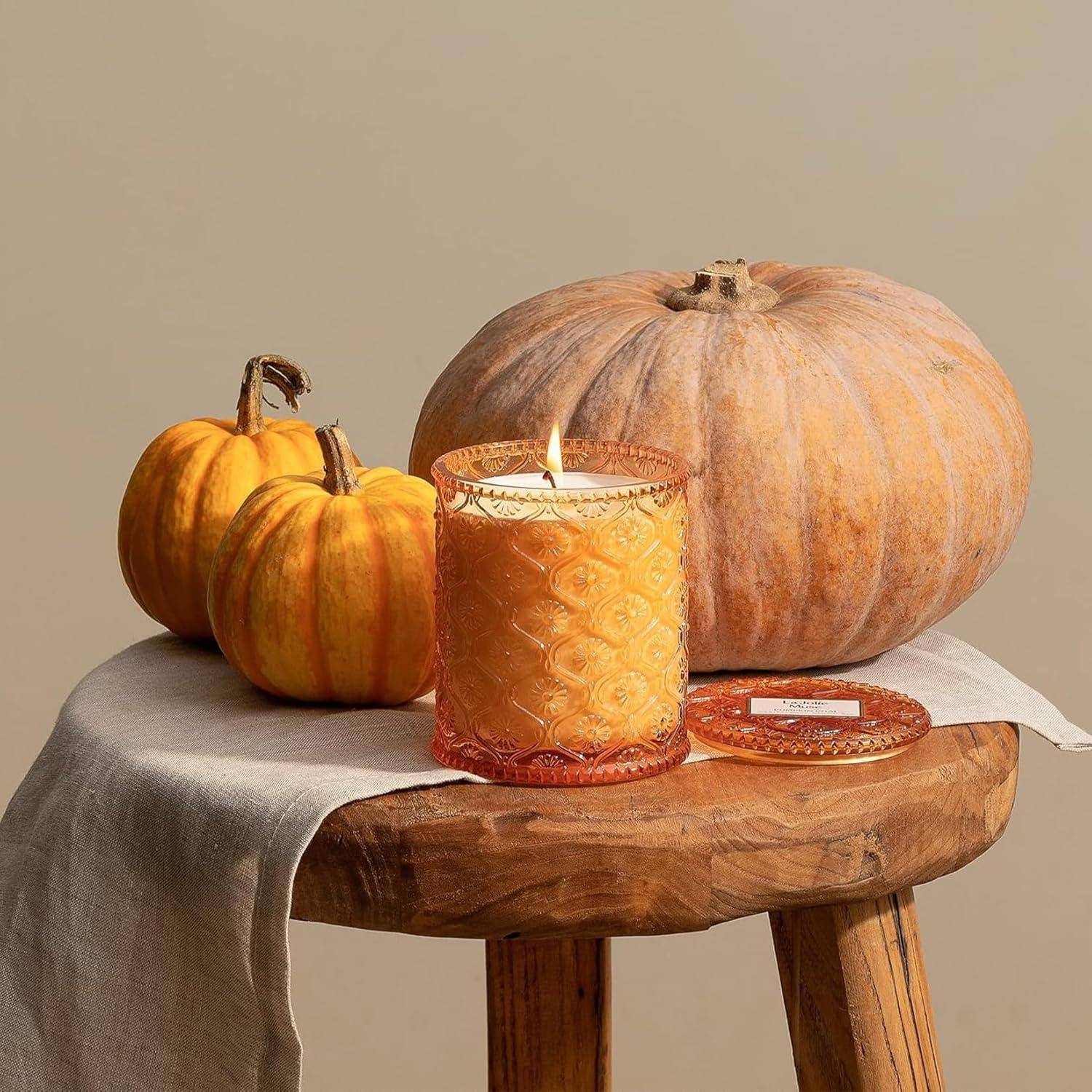Eine brennende Kerze auf einem Holztisch neben drei Kürbissen, die eine gemütliche, herbstliche Atmosphäre schaffen.
