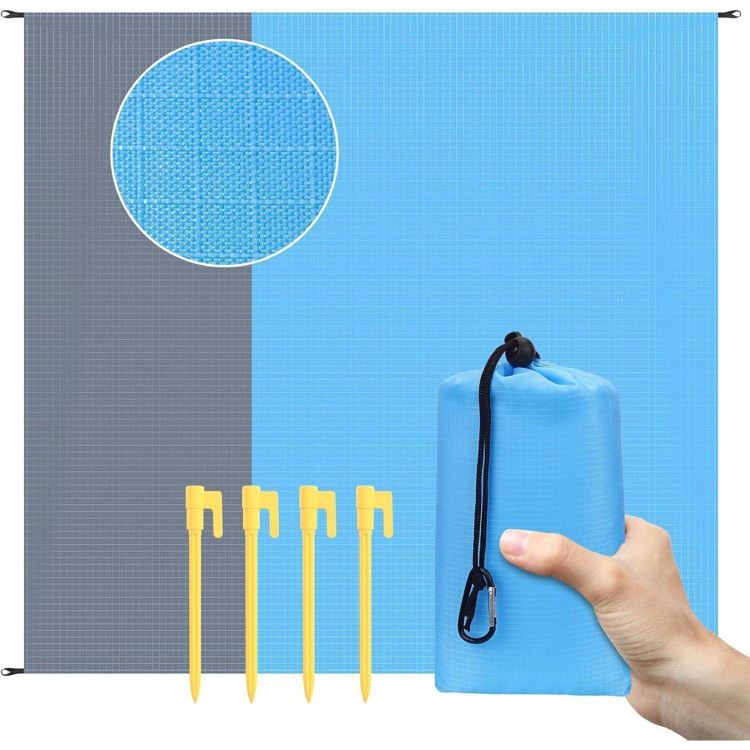 Eine Hand hält eine gefaltete blaue Strandmatte mit einem Karabiner. Die Matte ist teilweise geöffnet und zeigt ein zweifarbiges Blau- und Graumuster. Vier gelbe Heringe sind enthalten.