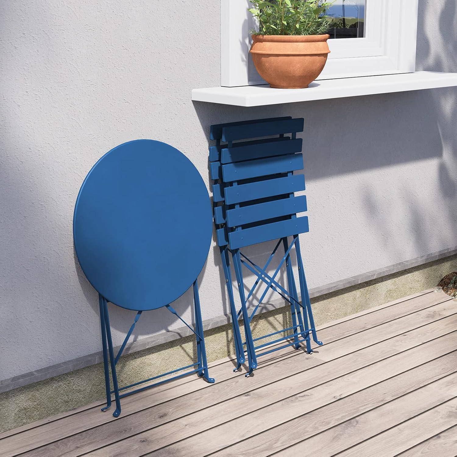 Blaue Klappstühle und ein runder Klapptisch sind ordentlich an einer Hauswand auf einer Holzterrasse gestapelt und für die Nutzung im Freien bereit.