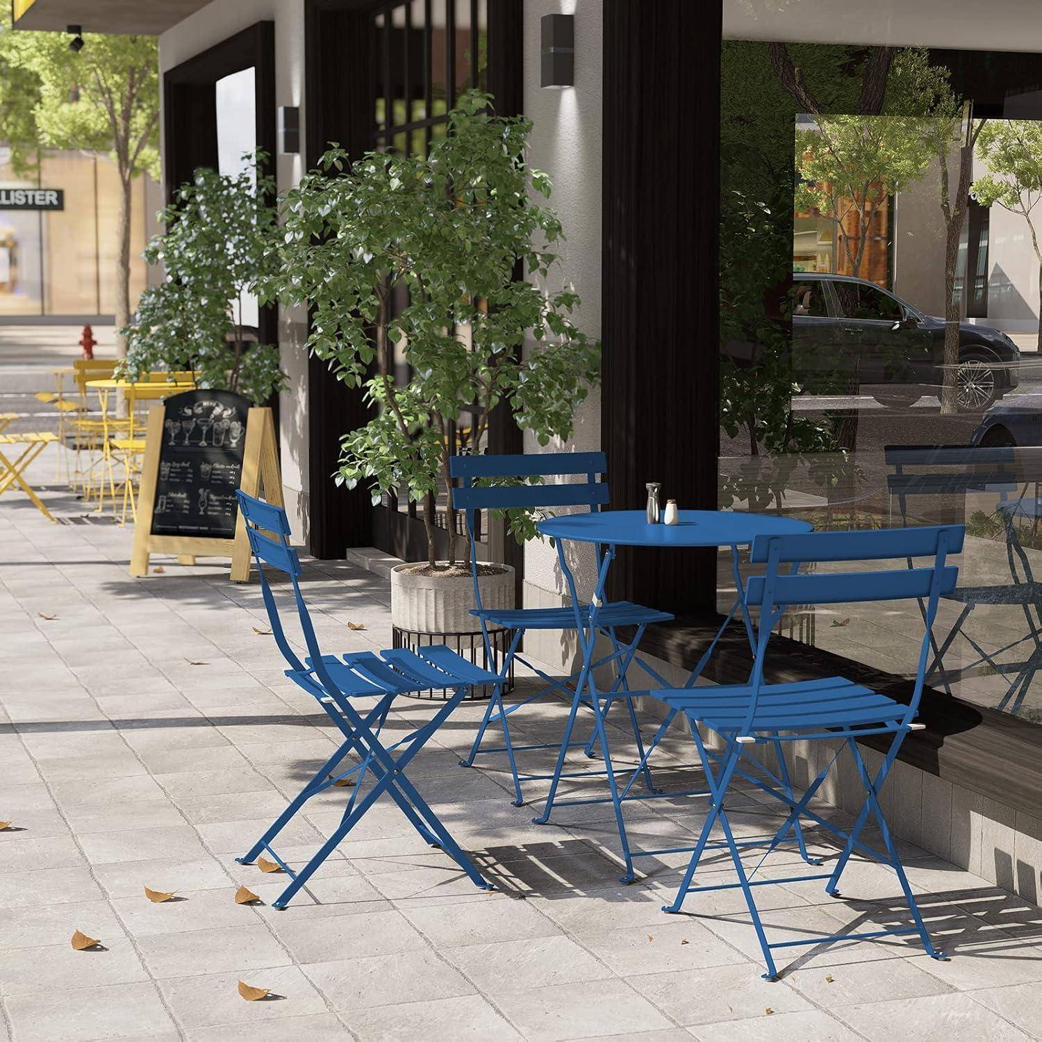 Eine Café-Terrasse mit blauen Tischen und Stühlen neben einem Geschäft, beschattet von Bäumen. Kreidetafeln zeigen Speisekarten; die Straße ist ruhig.
