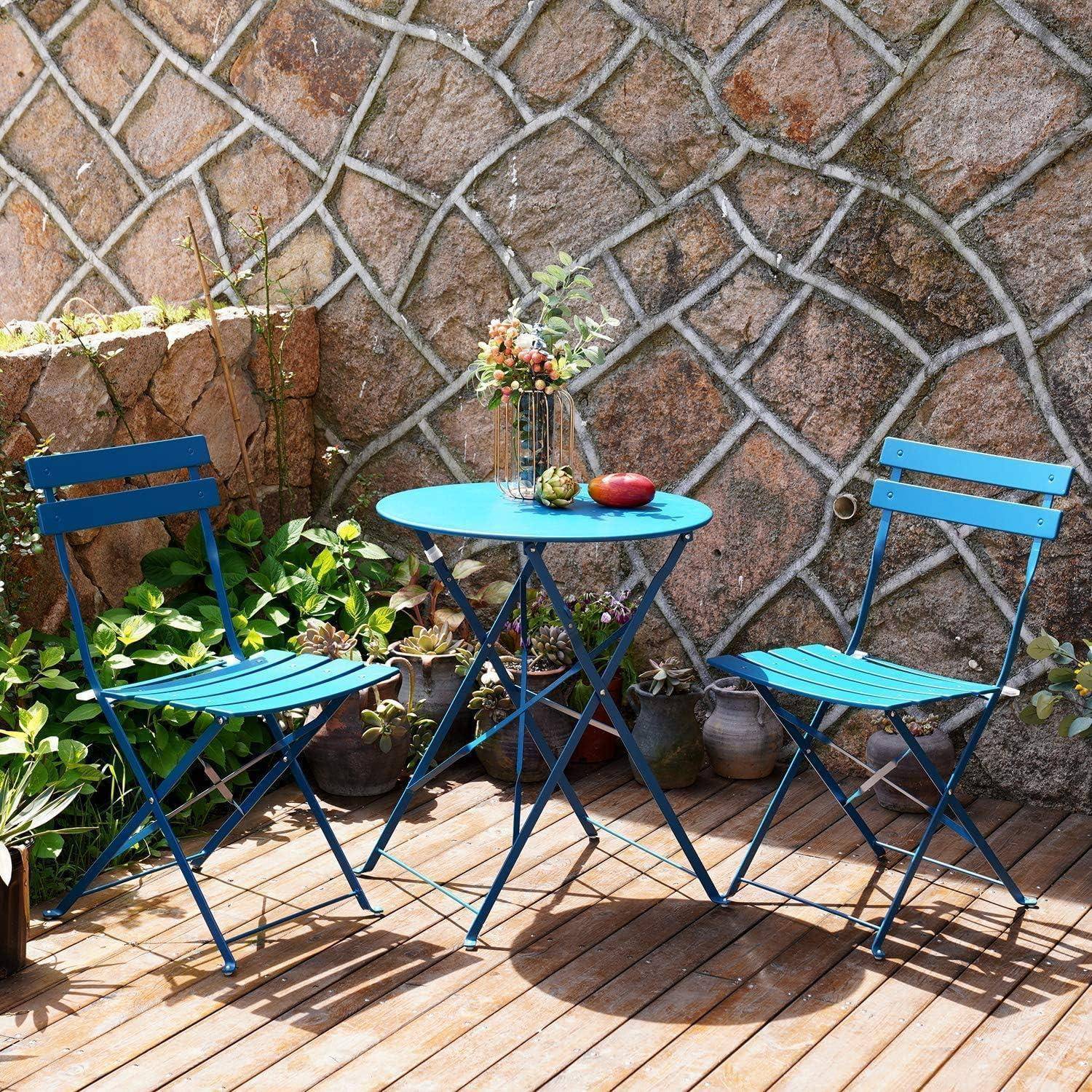 Zwei blaue Stühle und ein Tisch mit einer Vase voller Blumen und einem roten Apfel befinden sich auf einer Holzterrasse, vor einer strukturierten Steinwand im Hintergrund.