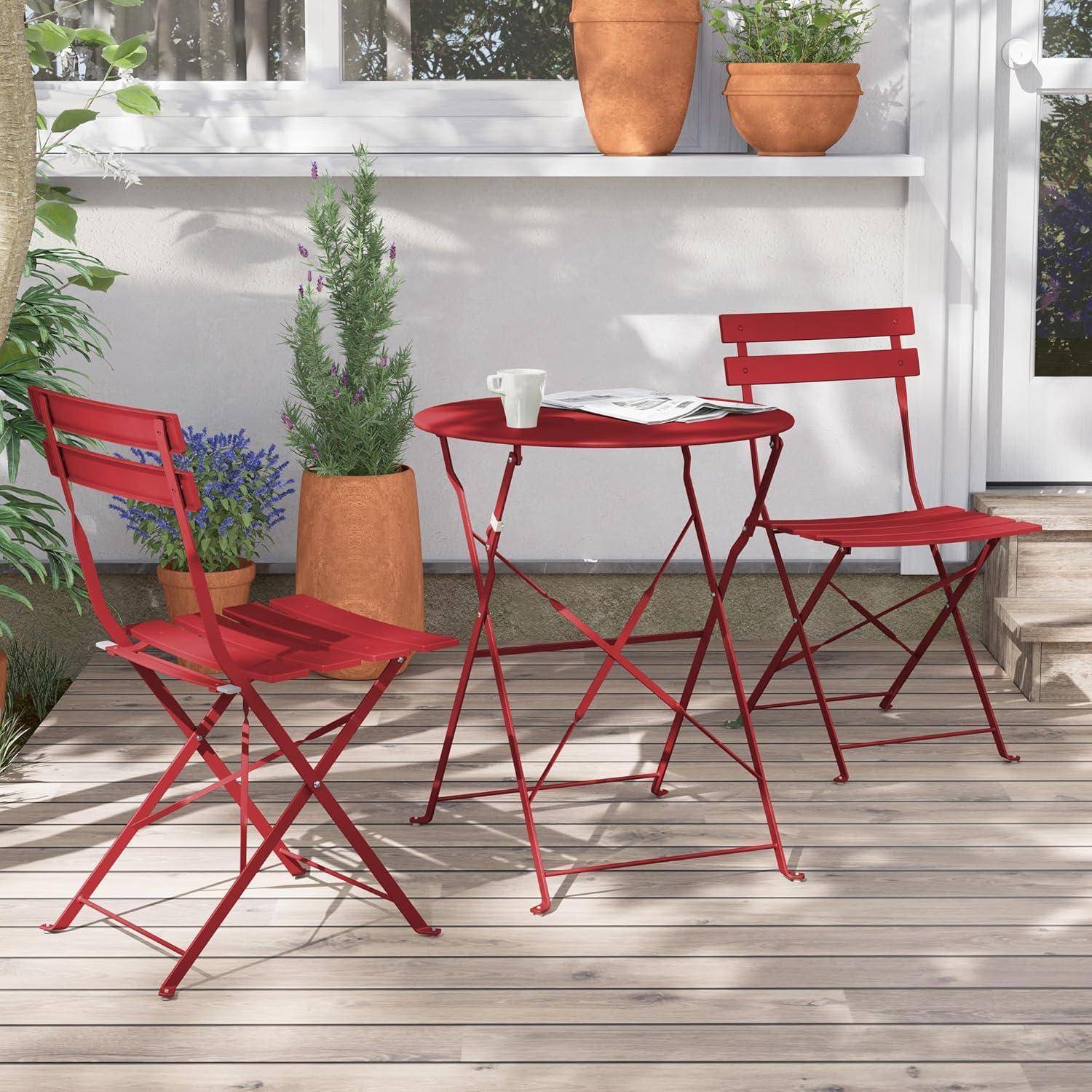 Ein roter Bistrotisch mit zwei roten Stühlen steht auf einer Holzterrasse. Eine Kaffeetasse steht auf dem Tisch. Topfpflanzen und Grünpflanzen umgeben die Szenerie.
