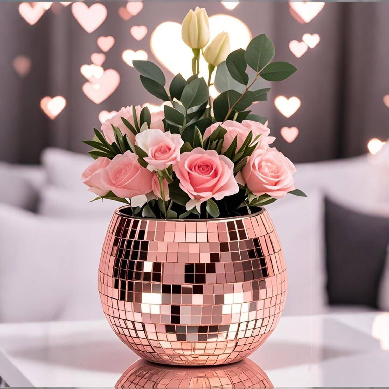 Ein Strauß rosa Rosen und eine gelbe Tulpe in einer reflektierenden, Discokugel-artigen Vase auf einem Glastisch; herzförmige Lichter im unscharfen Hintergrund.