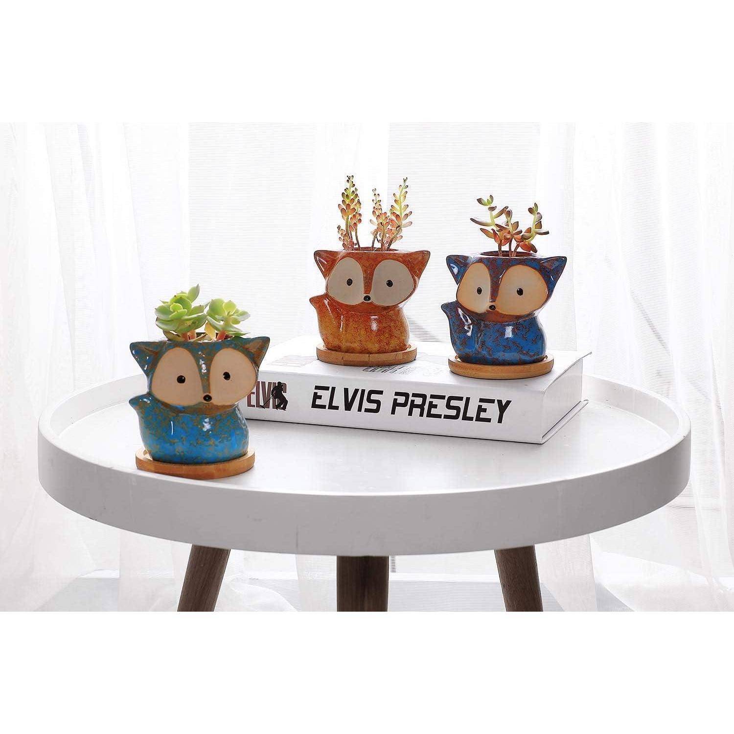 Drei fuchsförmige Pflanzentöpfe stehen auf einem runden weißen Tisch, jeder gefüllt mit üppiger Grünpflanze. Sie ruhen auf einem Buch mit dem Titel 