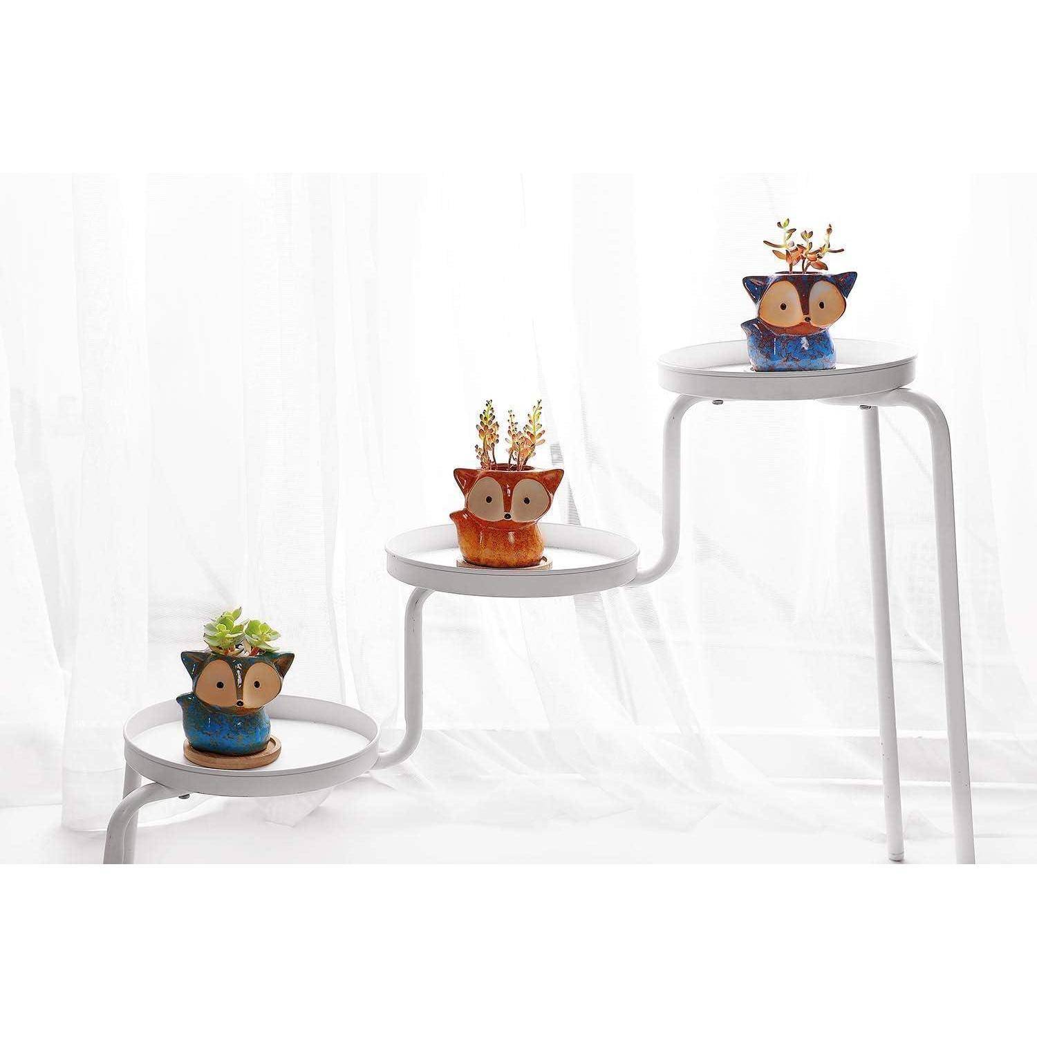 Drei kleine, tierförmige Pflanzgefäße mit Pflanzen stehen auf einem mehrstufigen weißen Ständer vor einem weißen Hintergrund.