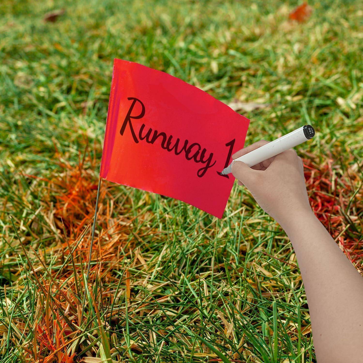 Eine Hand schreibt ‚Runway 1' auf eine kleine rote Fahne, die in grasbewachsenem Boden gepflanzt ist.