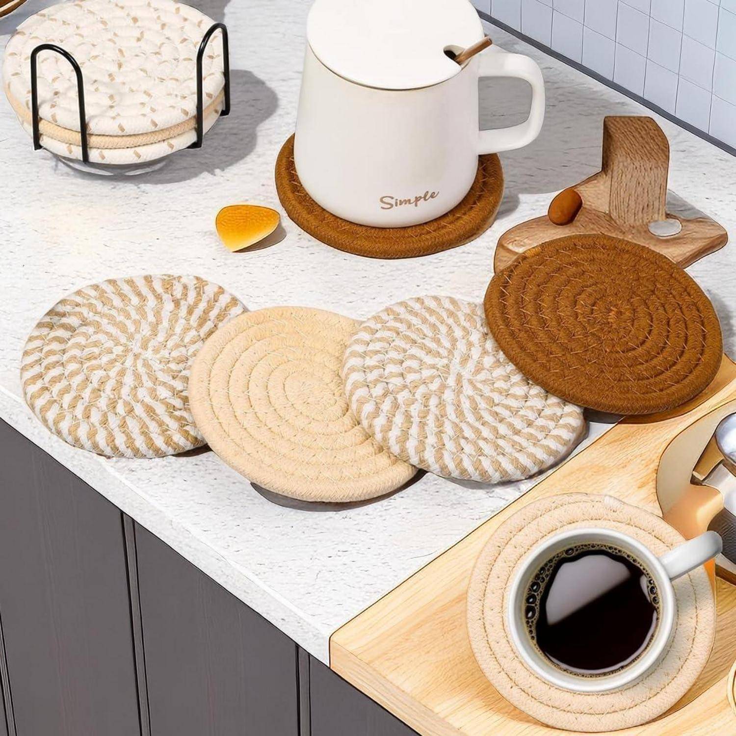 Eine Küchenarbeitsplatte mit einem weißen Wasserkocher auf einem Untersetzer, verschiedenen strukturierten Untersetzern, Keksen und einer Kaffeetasse auf einem Holztablett.