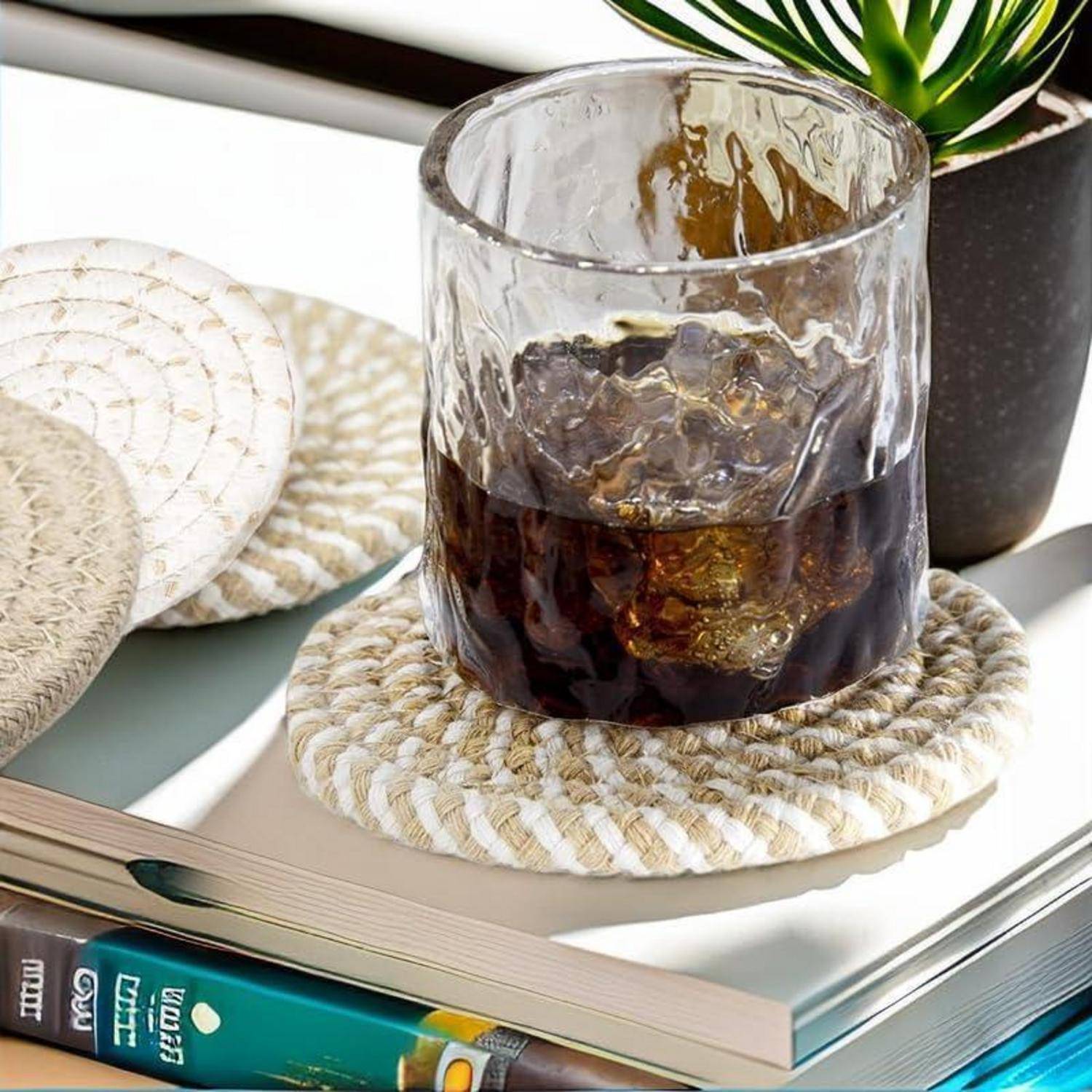 Ein Glas Eiskaffee auf einem Untersetzer neben einem geschlossenen Buch und einer kleinen Topfpflanze auf einer reflektierenden Oberfläche.