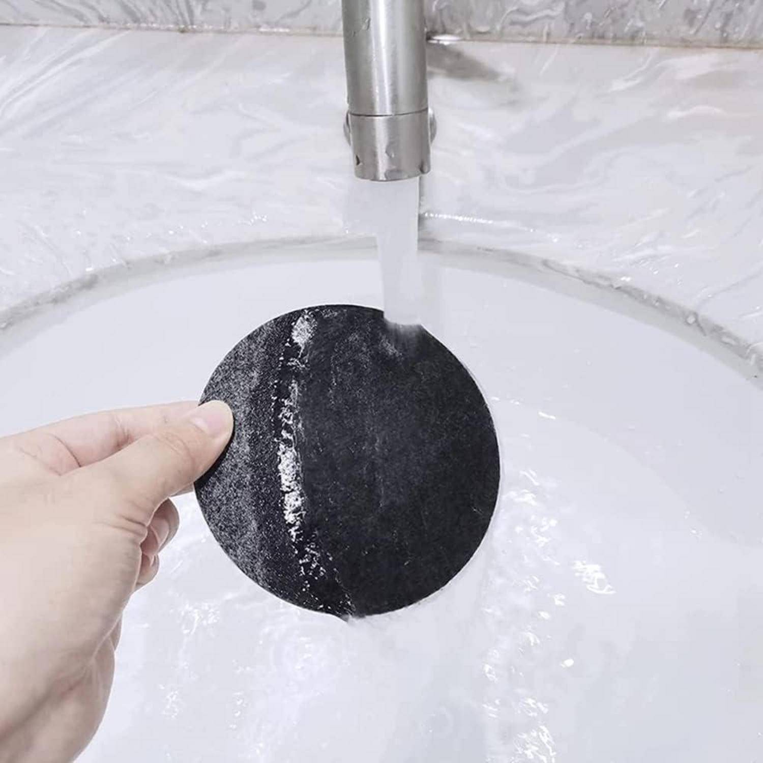 Eine Hand, die einen schwarzen Schwamm unter einem laufenden Wasserhahn in einem Waschbecken hält und den Schwamm abspült oder reinigt.