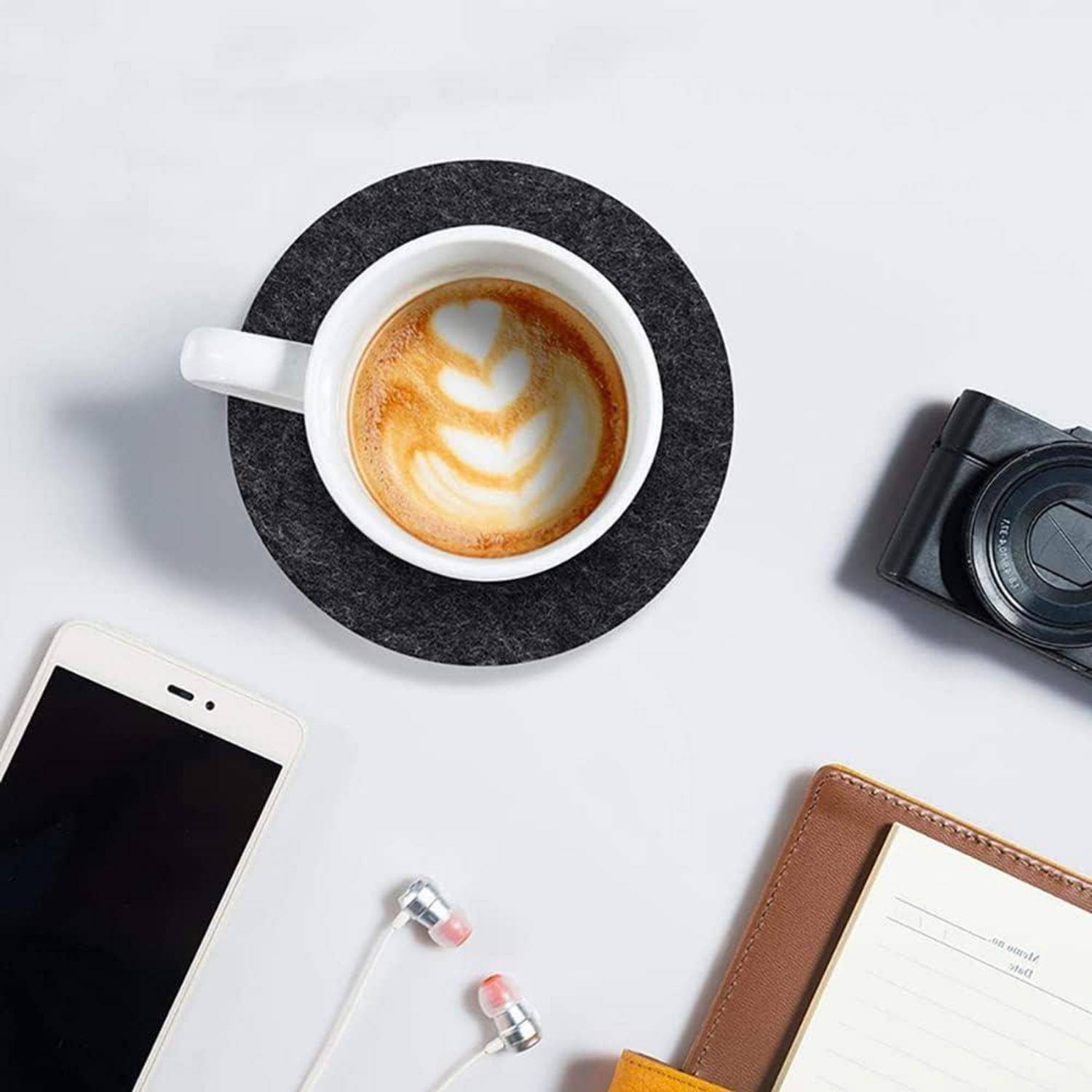 Eine Draufsicht eines Cappuccinos mit Latte Art, ein Smartphone mit Kopfhörern, ein Notizbuch und eine Kamera auf einer weißen Oberfläche.