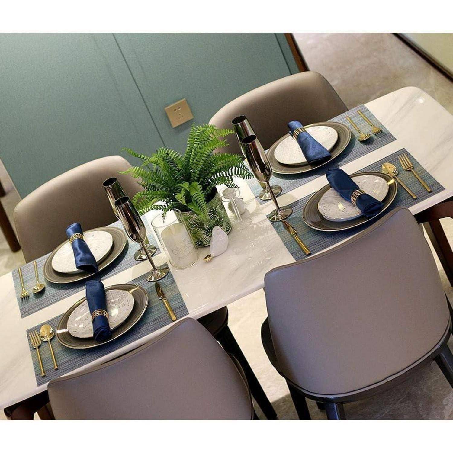 Eleganter Esstisch für vier Personen, ausgestattet mit blauen Platzdeckchen, goldenem Besteck und marineblau-farbenen Servietten. Ein grünes Pflanzenmittelstück ergänzt die Dekoration.