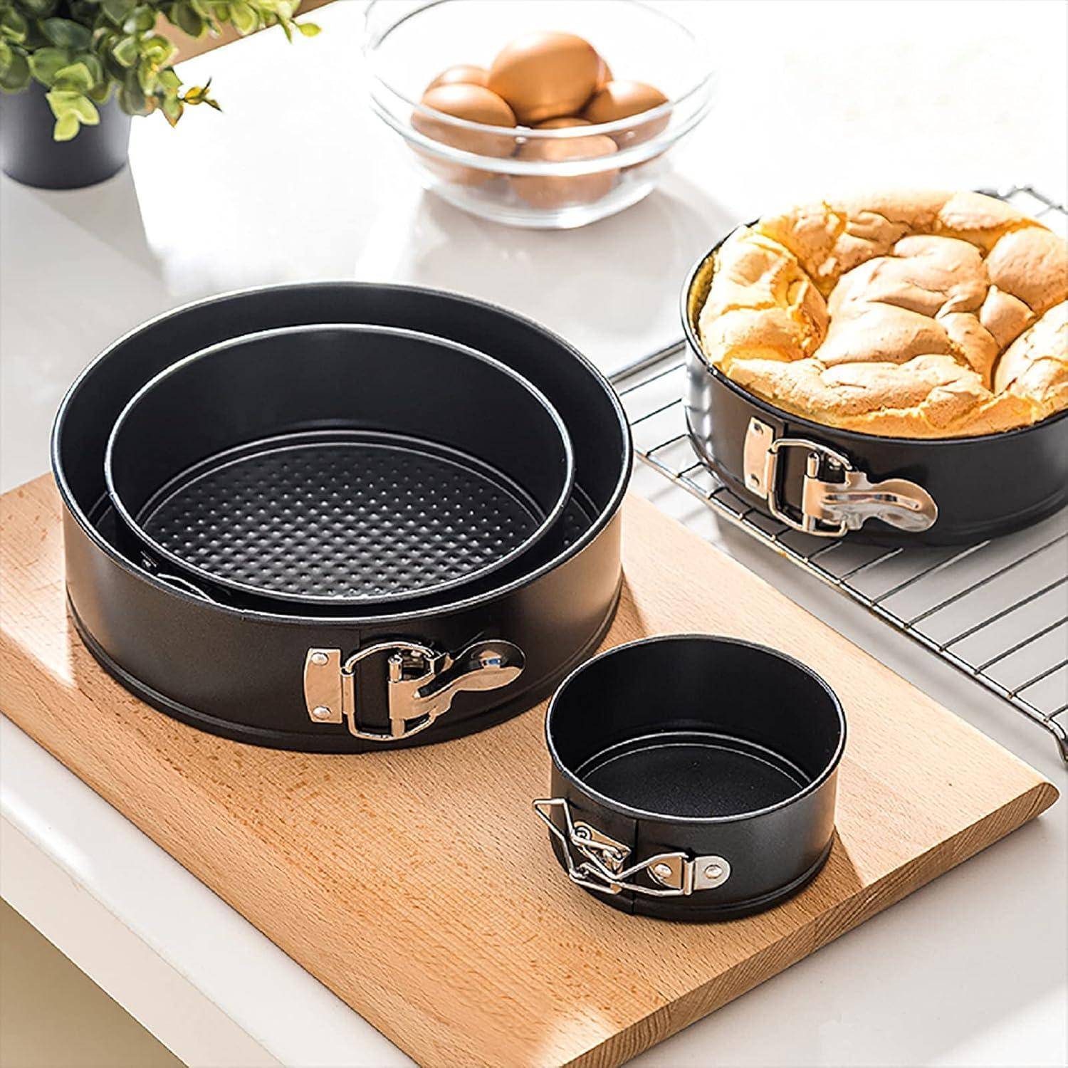Ein Satz schwarze antihaftbeschichtete Springformen in verschiedenen Größen auf einem Holzbrett, daneben ein frisch gebackener Kuchen, der auf einem Kuchengitter abkühlt.