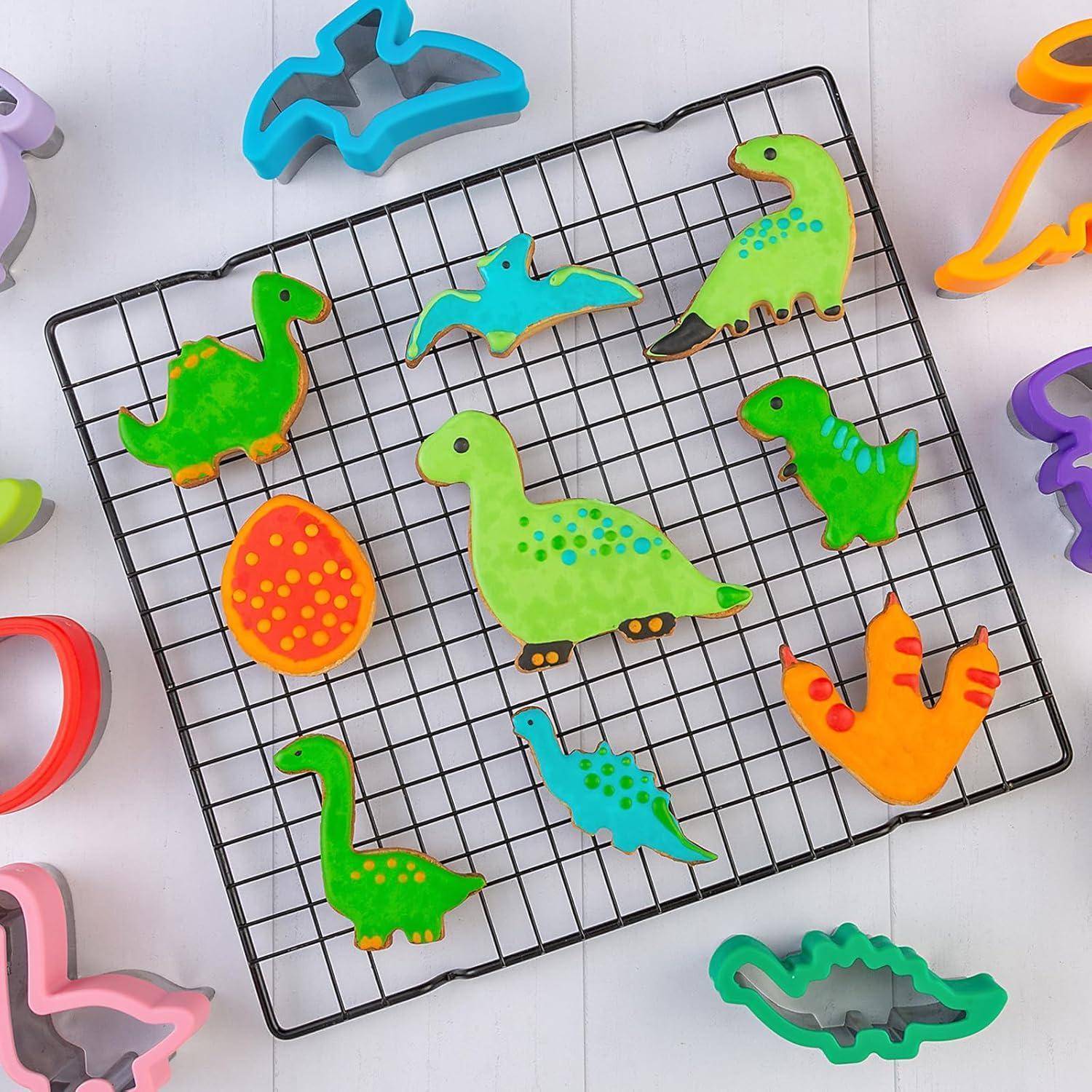 Dinosaurier-Plätzchen in verschiedenen Formen und Farben, darunter grüne Dinosaurier und ein geflecktes Ei, ausgelegt auf einem Kuchengitter mit Ausstechformen.