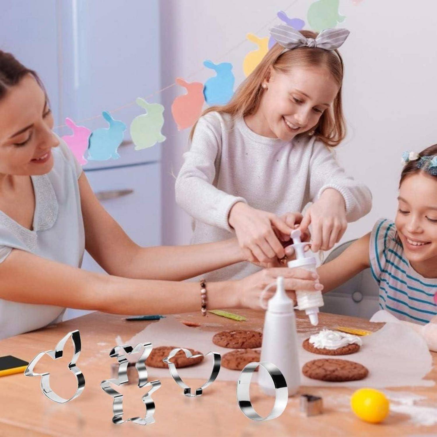 Eine Frau und zwei Kinder dekorieren Plätzchen an einem Tisch mit Ausstechformen und Zuckerguss, lächeln und beschäftigen sich gemeinsam mit einer lustigen Aktivität.
