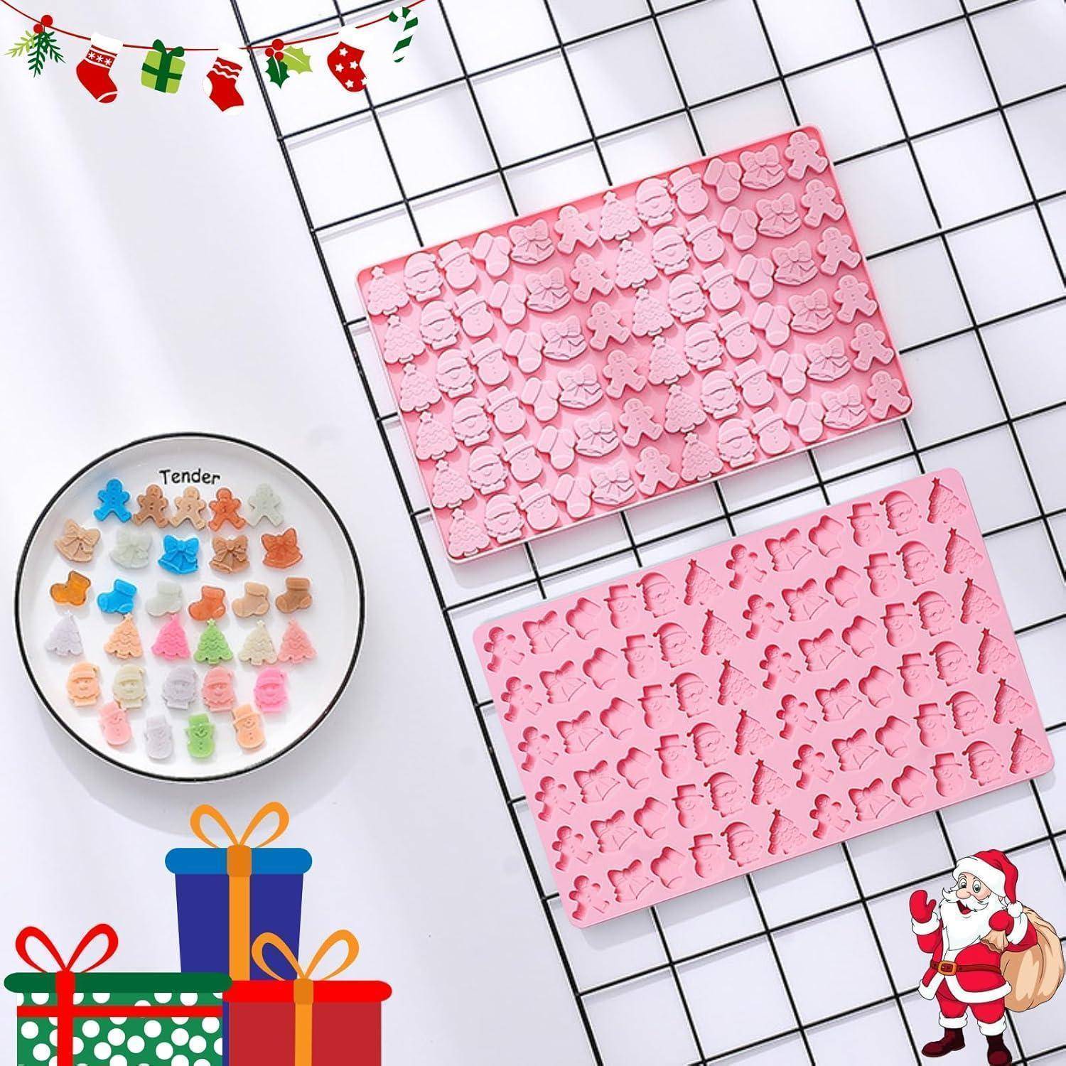 Zwei rosa Silikonformen mit Weihnachtsformen wie Bäumen und Schneemännern, neben einem Teller mit bunten Gummibärchen. Weihnachtliche Dekoration umgibt sie.