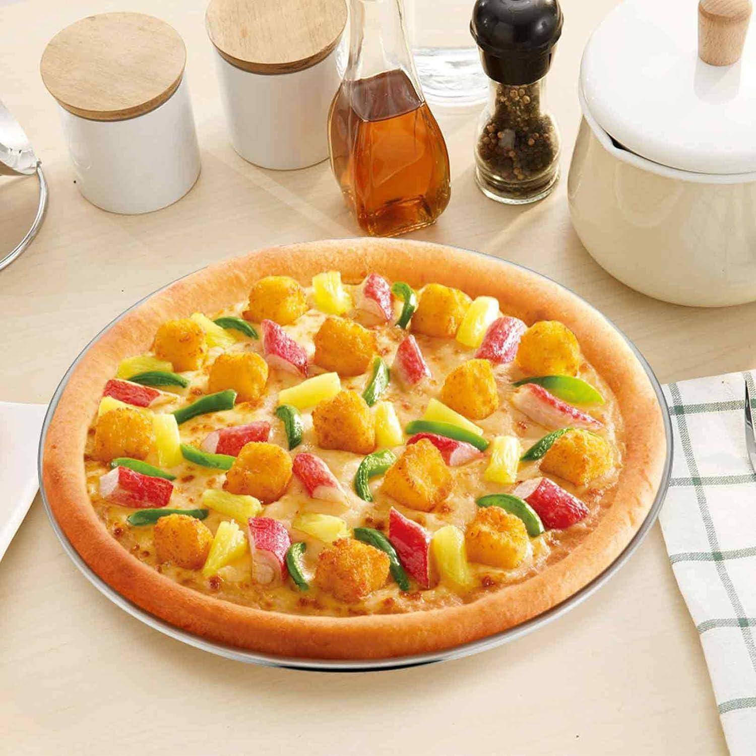 Eine Pizza belegt mit Ananas, Surimi-Sticks und grünen Paprikaschoten auf einem Tisch mit Gewürzen und einer karierten Serviette.