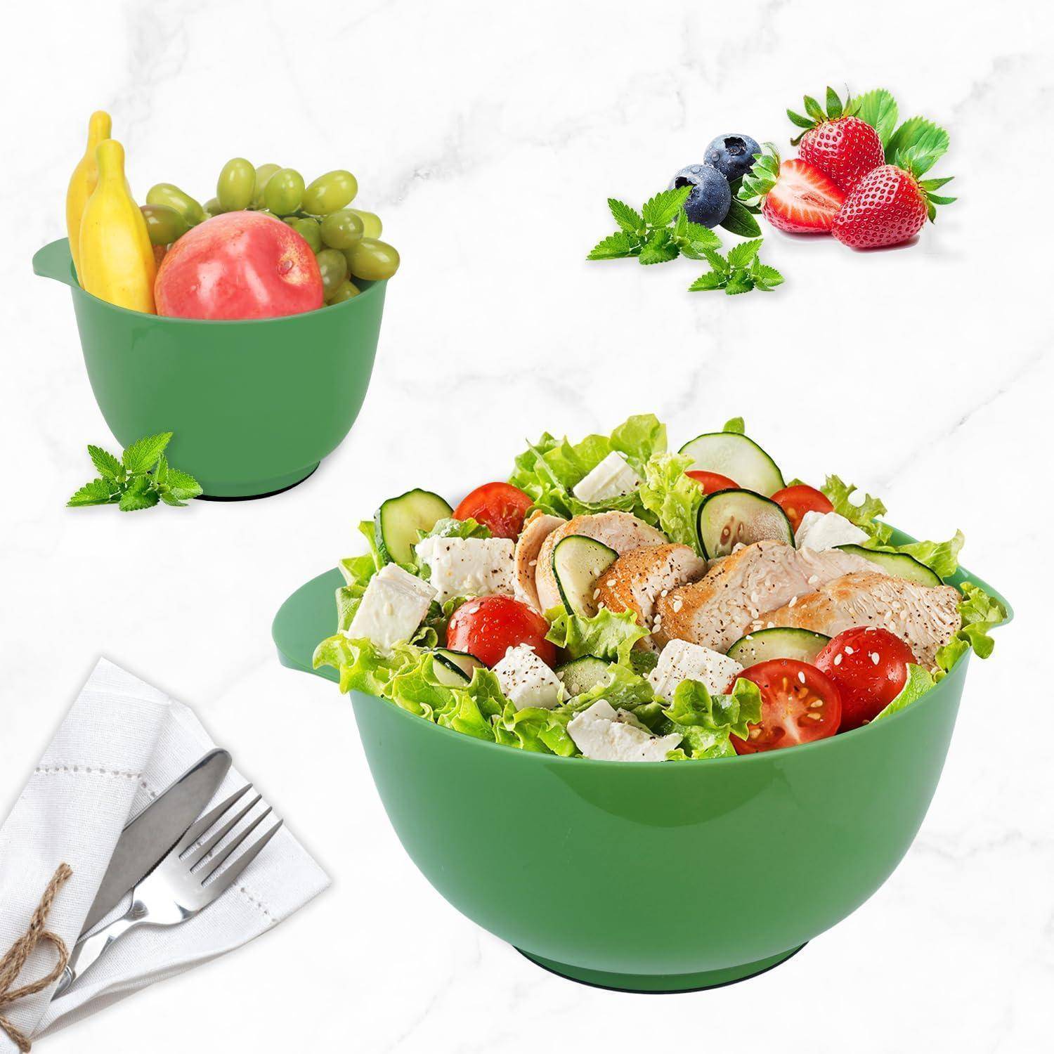 Eine grüne Schüssel mit einem frischen Salat, der Salat, Tomaten, Gurken, Hühnchen und Käse enthält. Eine kleinere Schüssel mit Obst (Banane, Apfel, Trauben) befindet sich im Hintergrund. Eine Gabel und ein Messer sind in einer Serviette gewickelt in der Nähe.