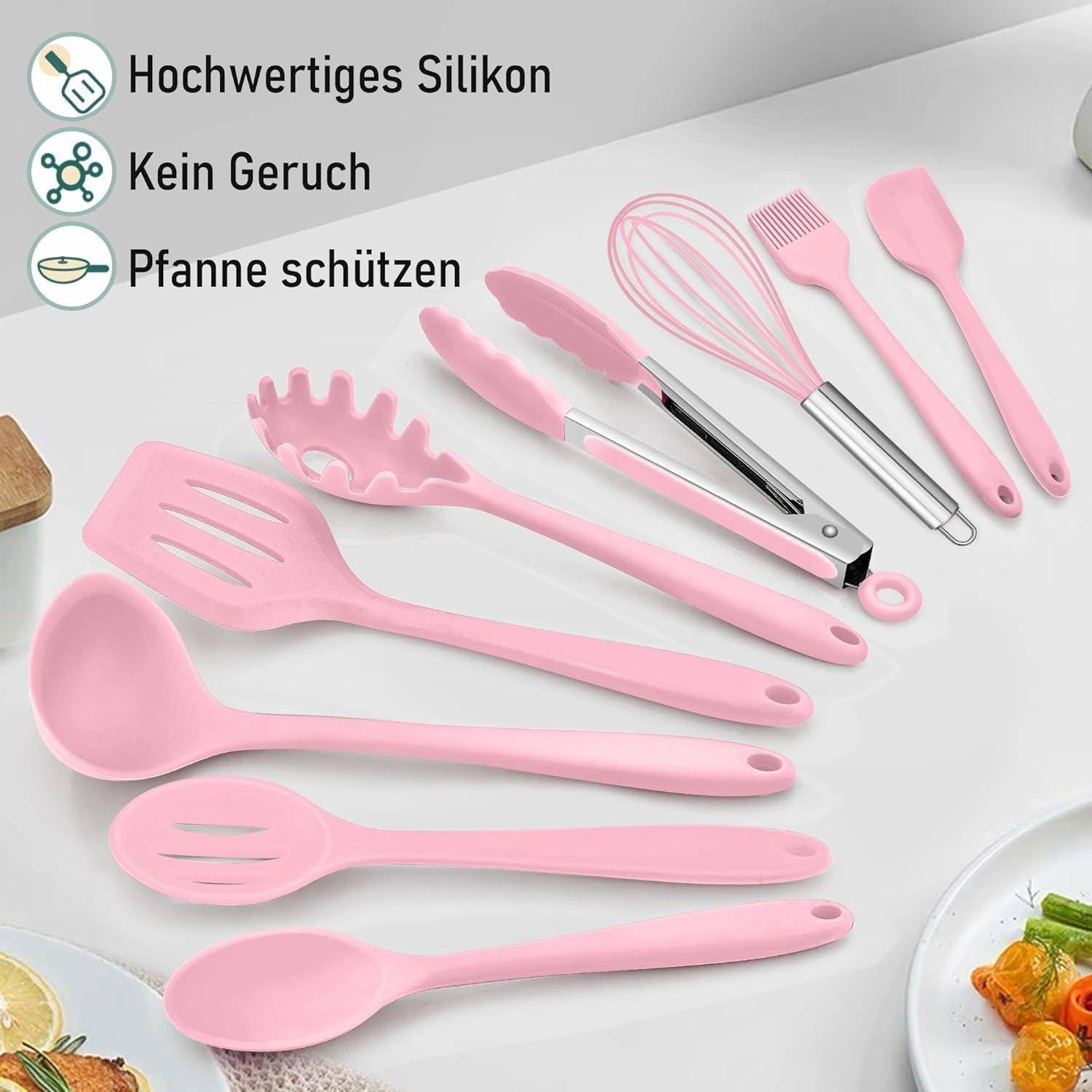 Ein Set pinker Silikonküchenutensilien, bestehend aus Löffeln, Pfannenwender, Zange, Schneebesen und Pinsel, auf einer Arbeitsplatte neben einem Teller.