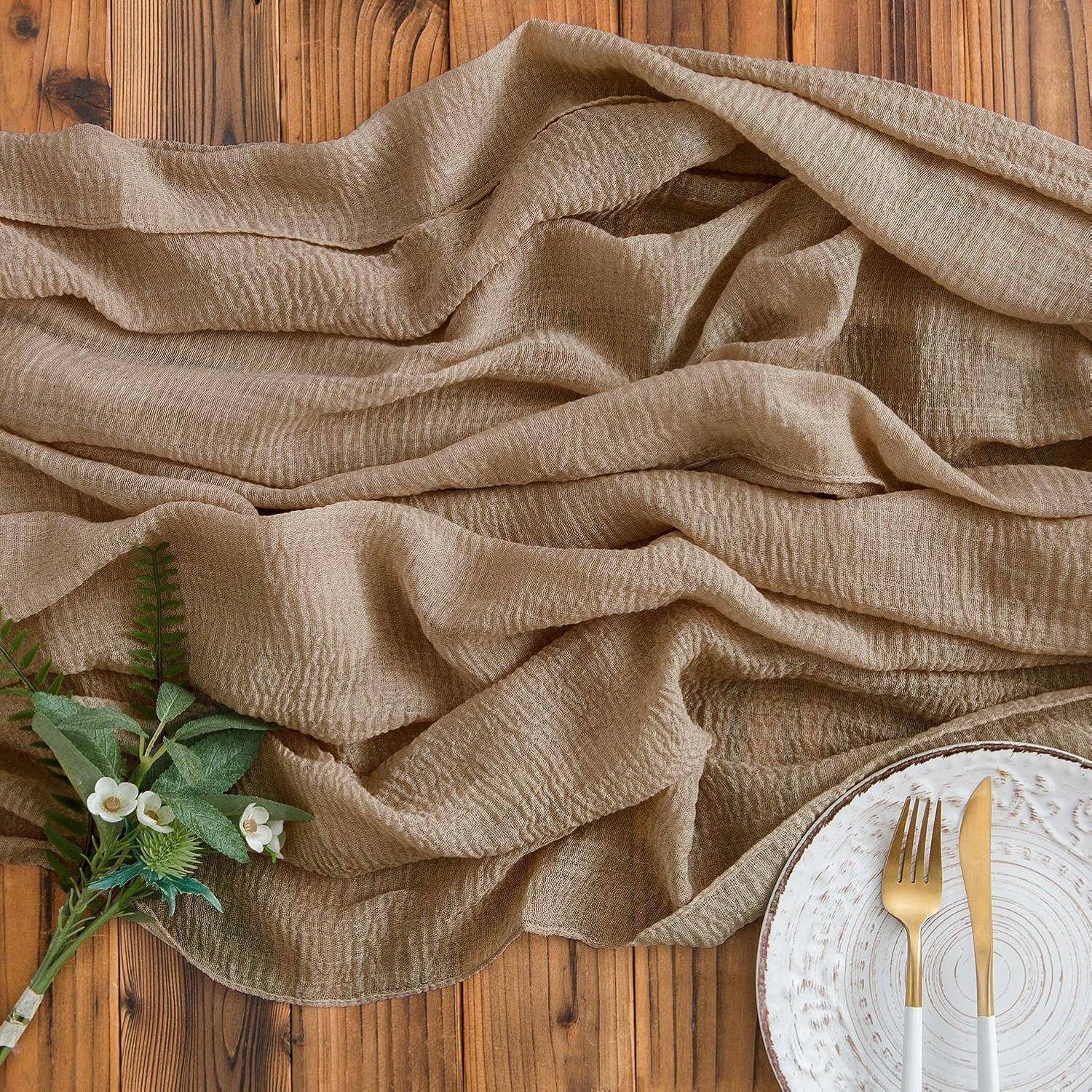 Ein zerknittertes beiges Tuch ist über einen Holztisch ausgebreitet. Darauf ein dekorativer Zweig mit grünem Laub und weißen Blüten sowie ein Teller mit goldenen Besteck.