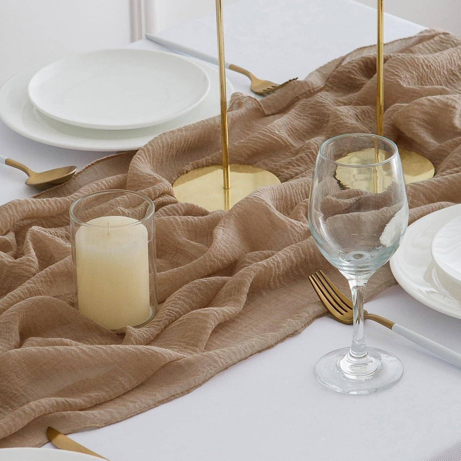 Eine minimalistische Tischdeckung besteht aus einer weißen Tischdecke mit einem beigen Tischläufer, goldenen Kerzenhaltern, einer Kerze und Glasgeschirr.