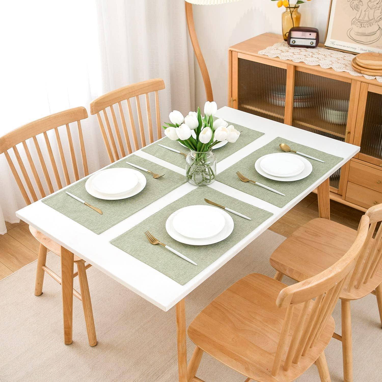 Ein Tisch für vier Personen gedeckt mit grünen Tischsets, weißen Tellern, Besteck und einer Vase mit weißen Blumen in der Mitte. Holzstühle umgeben ihn.