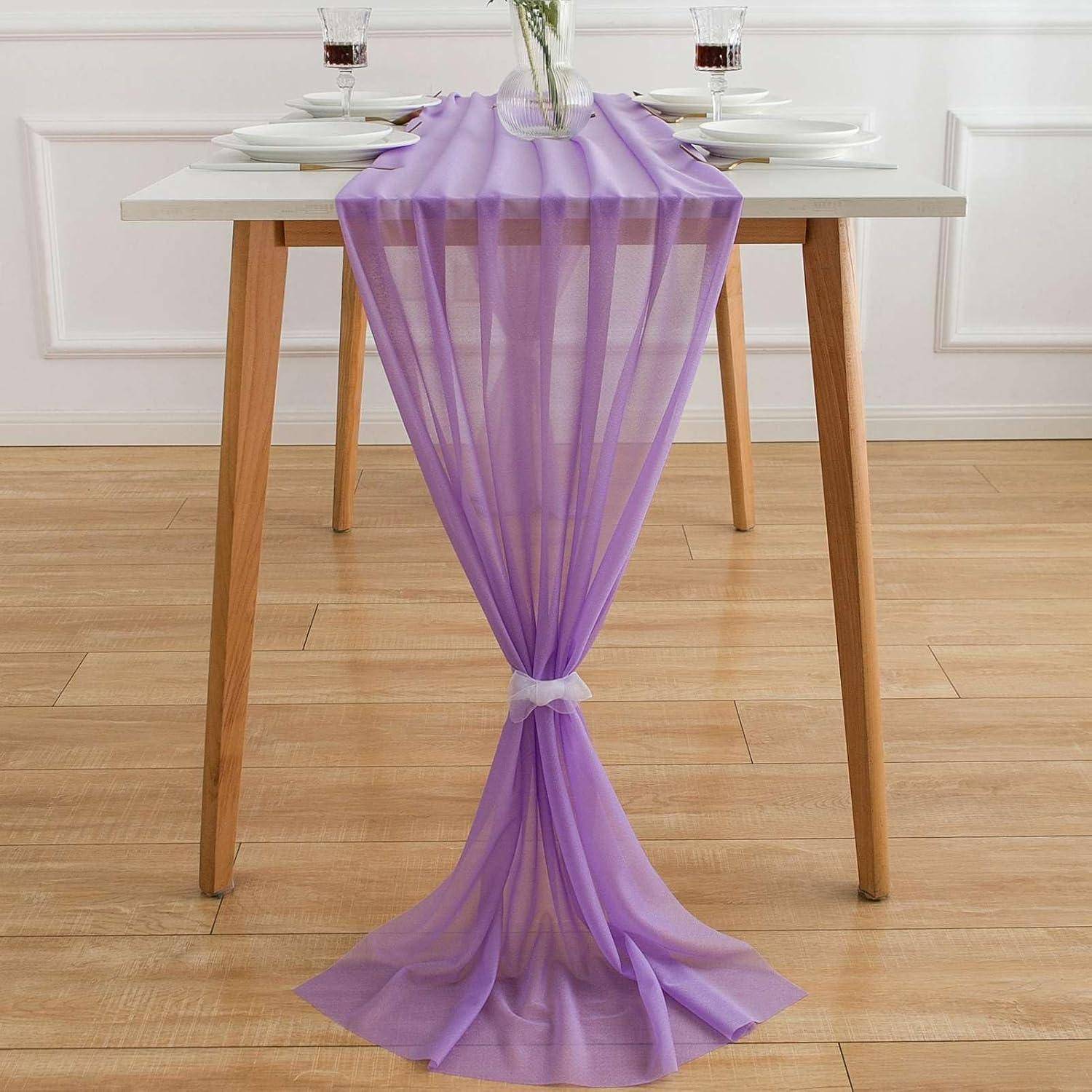 Ein weißer Tisch mit Holzbeinen ist mit einem durchsichtigen lila Tischläufer geschmückt, der sich elegant bis zum Boden erstreckt und in der Mitte mit einer Bindung zusammengehalten wird.