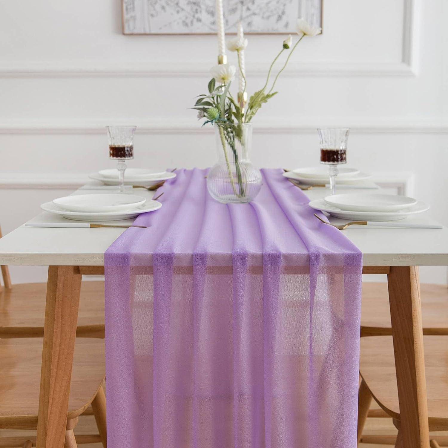 Ein für vier Personen gedeckter Tisch mit eleganten weißen Tellern, dekorativen Gläsern und einem Lavendel-Tischläufer. Ein Blumengesteck mit Kerzen ist vorhanden.