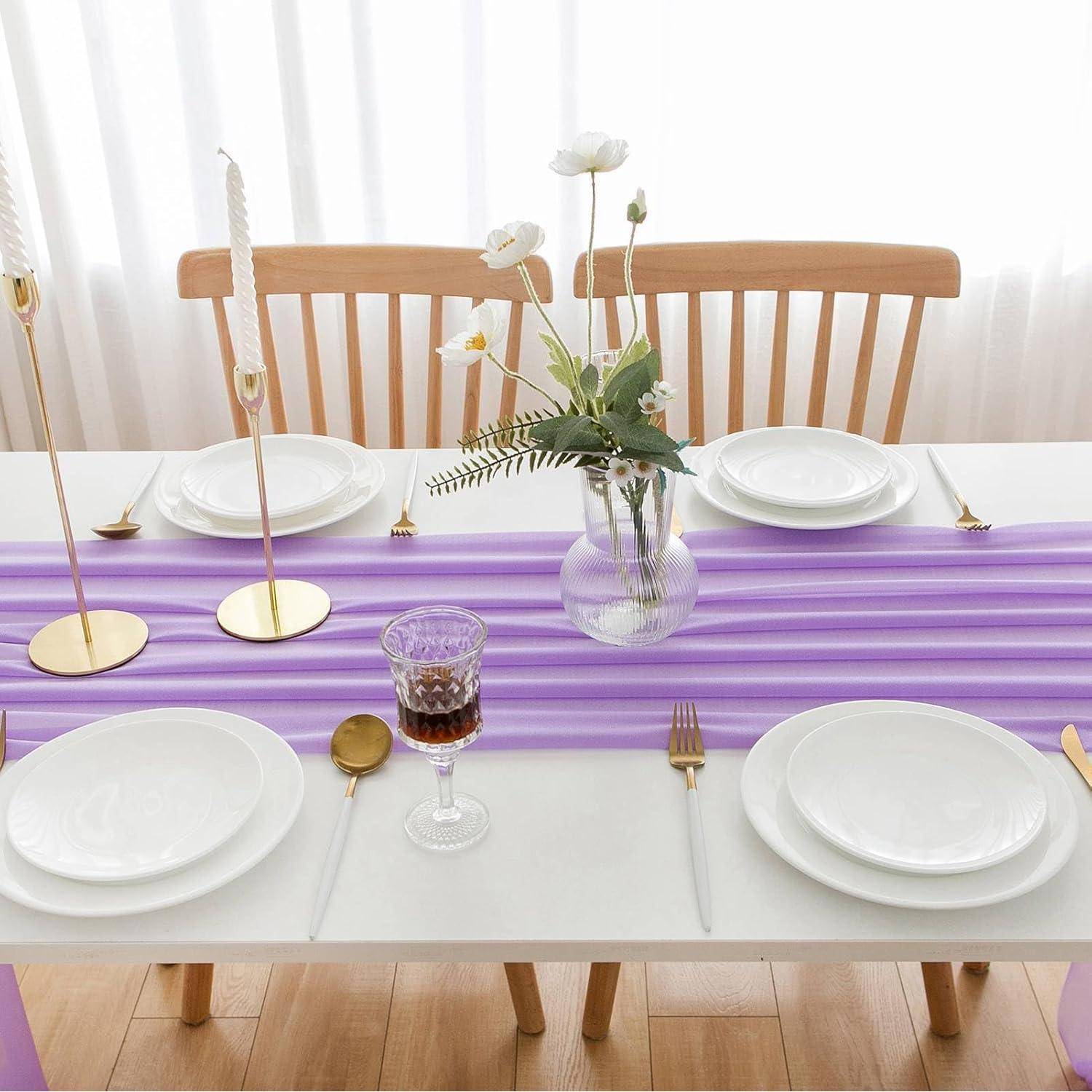 Ein Esstisch für vier Personen, gedeckt mit weißen Tellern, goldenen Bestecken, einem lila Tischläufer und einer Blumenvase als Mittelpunkt. Leichte Gardinen und Kerzen vervollständigen das Ambiente.