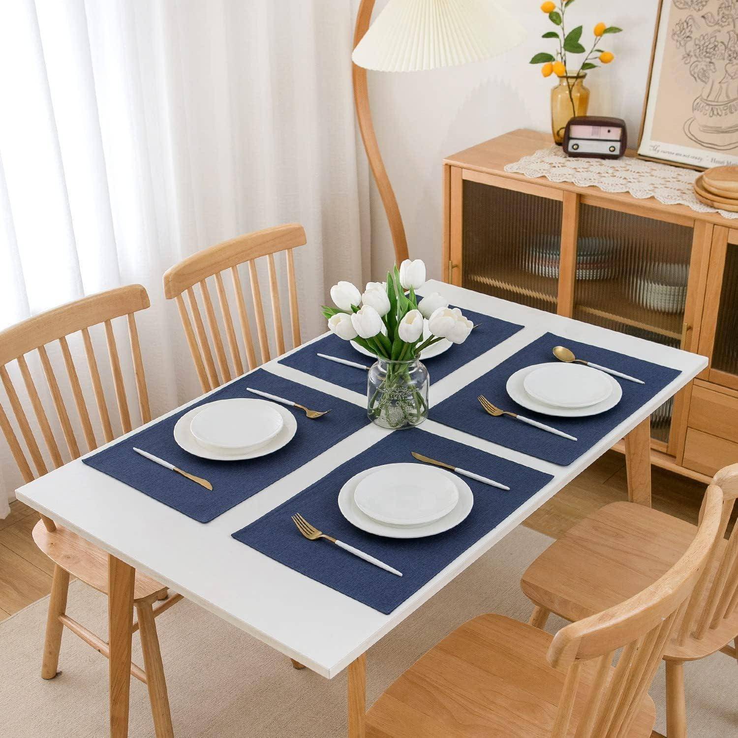 Ein Esstisch, gedeckt für vier Personen, mit weißen Tellern auf marineblau-farbenen Platzsets, goldenem Besteck und einer Vase mit weißen Blumen. Helle Holzstühle umgeben den Tisch.