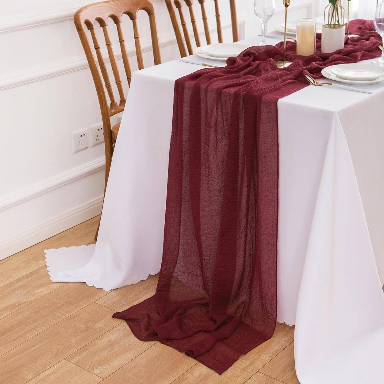 Ein Esstisch mit einer weißen Tischdecke, die mit einem marronefarbenen Stoff drapiert ist, ist für eine Mahlzeit gedeckt. Holzstühle umgeben den Tisch.