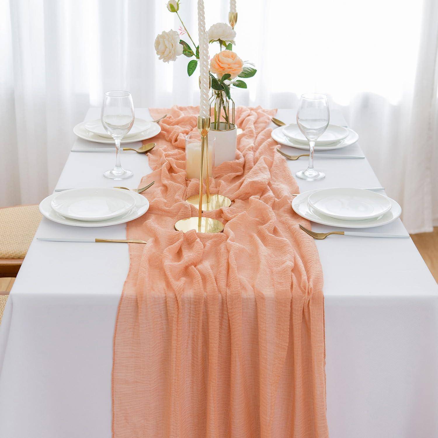 Ein Tisch für vier Personen gedeckt mit weißen Tellern, goldenen Bestecken und einem dekorativen pfirsichfarbenen Läufer mit einem Blumengesteck als Mittelpunkt.