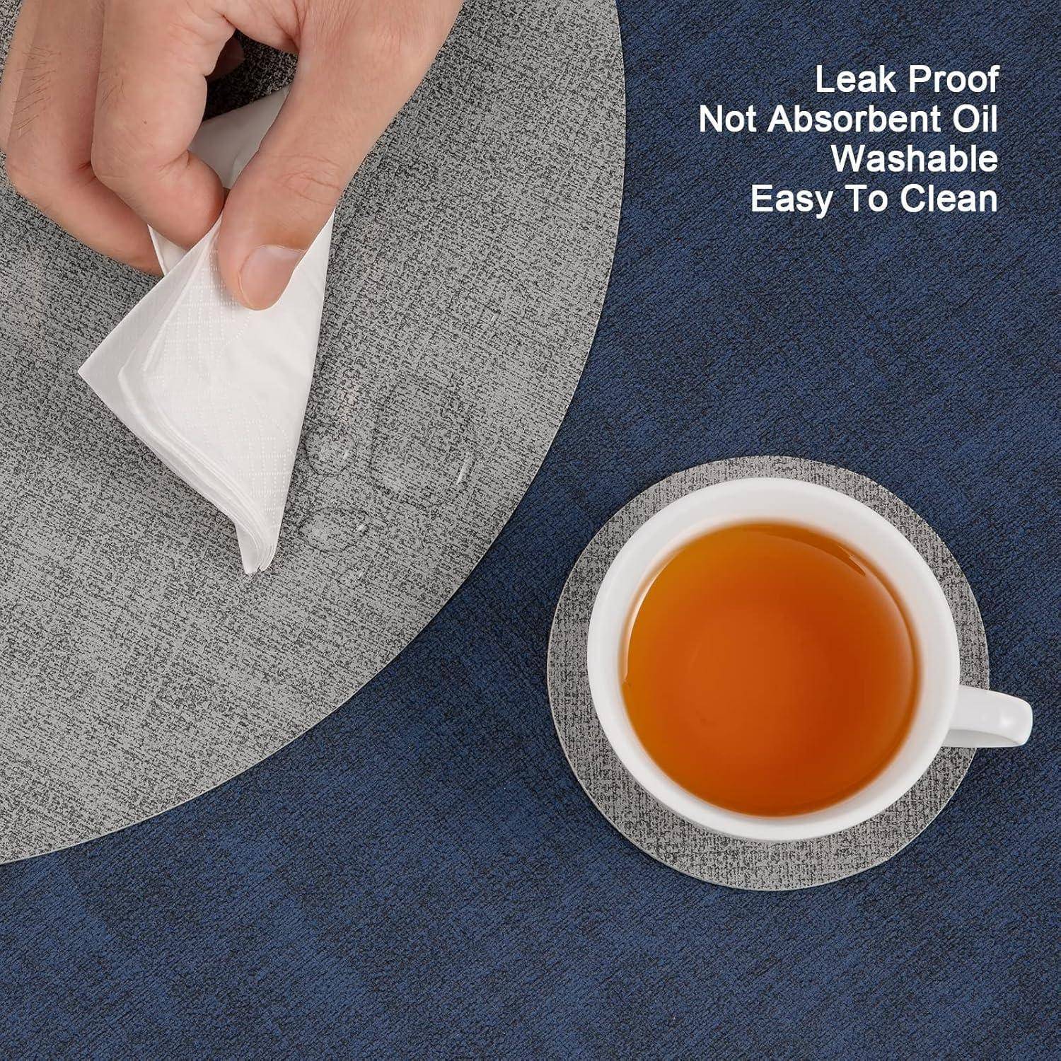 Eine Hand präsentiert eine weiße Serviette auf einer grauen, strukturierten, auslaufsicheren Platzunterlage neben einer weißen Teetasse auf einem passenden Untersetzer. Text lautet: ‚Auslaufsicher, Nicht Öl-Absorbierend, Waschbar, Leicht Zu Reinigen'.