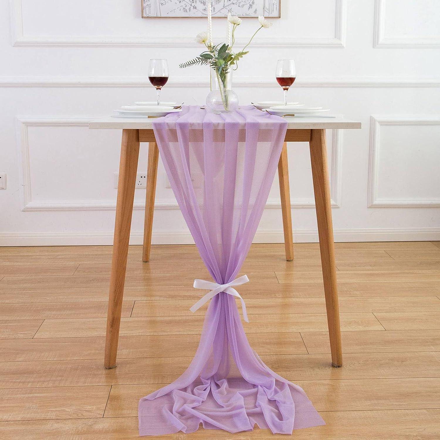 Ein Esstisch mit einem lila Tischläufer, zwei Weingläsern und einer weißen Vase mit Blumen. Zwei Stühle sind im Hintergrund teilweise sichtbar.