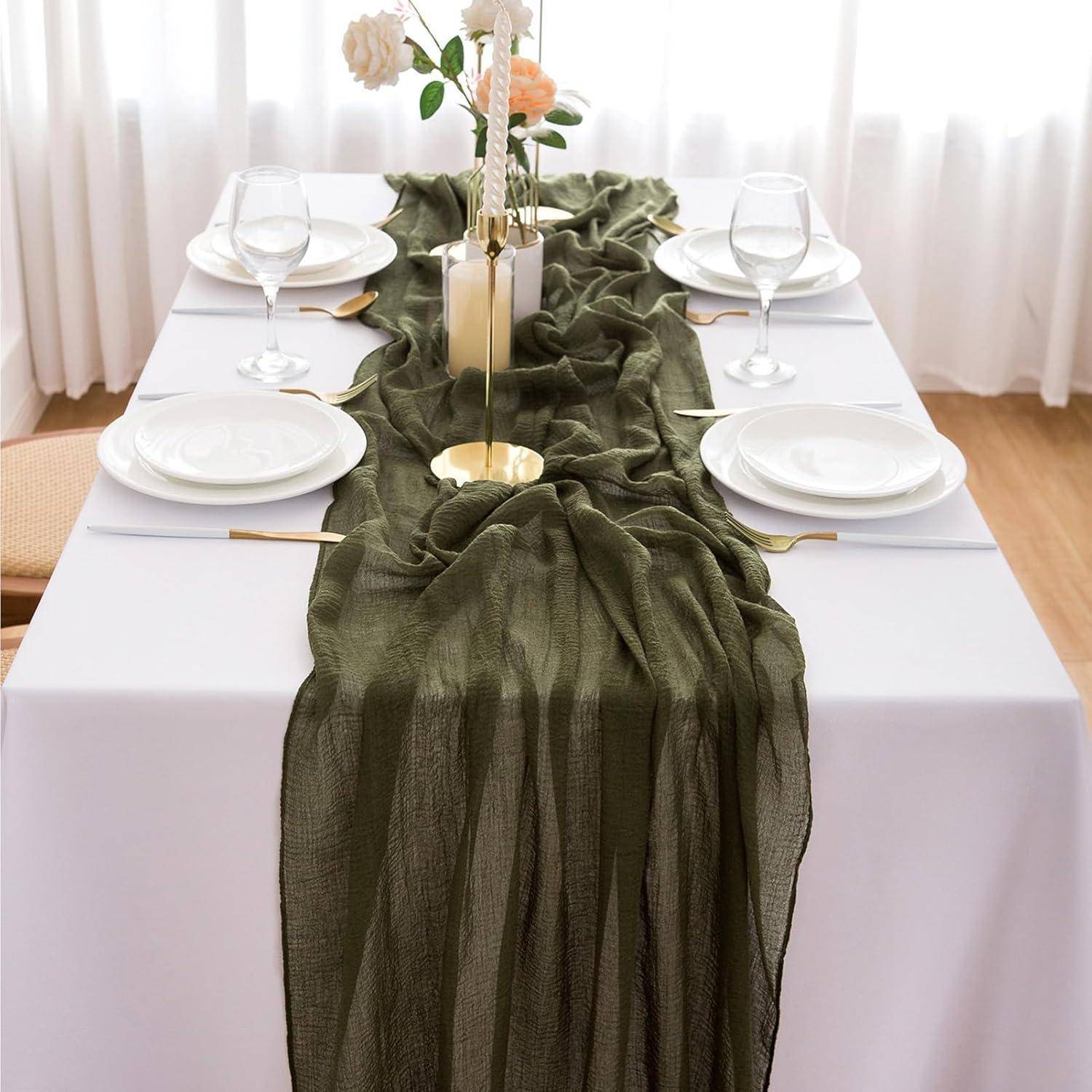 Ein Esstisch für vier Personen, gedeckt mit einer weißen Tischdecke, einem olivgrünen Tischläufer, weißen Tellern, Weingläsern und einem Blumengesteck in der Mitte.