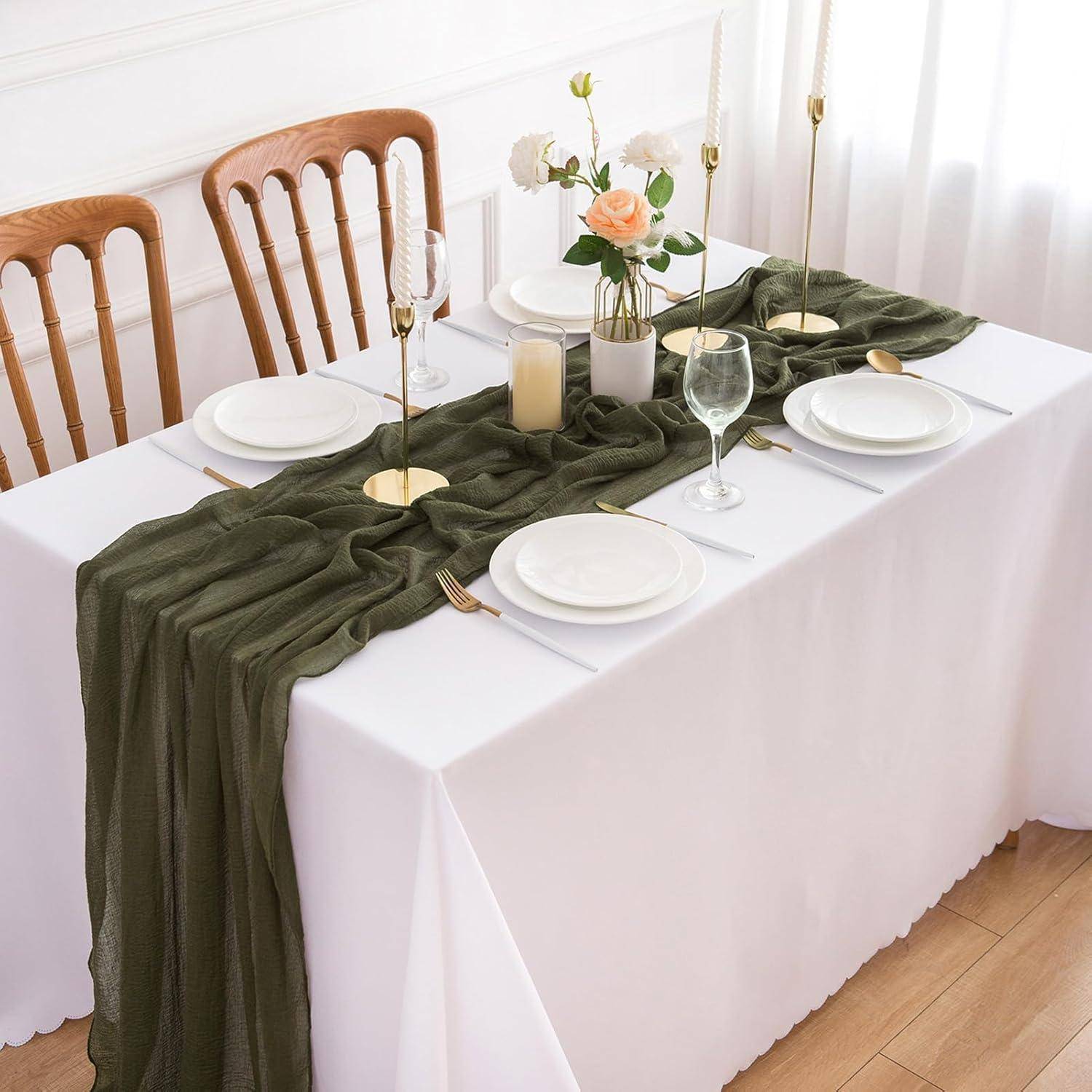 Ein Esstisch, der mit weißen Tellern, goldenem Besteck und einem dunkelgrünen Tischläufer gedeckt ist. Kerzenständer und eine Blumenarrangement verleihen Eleganz.