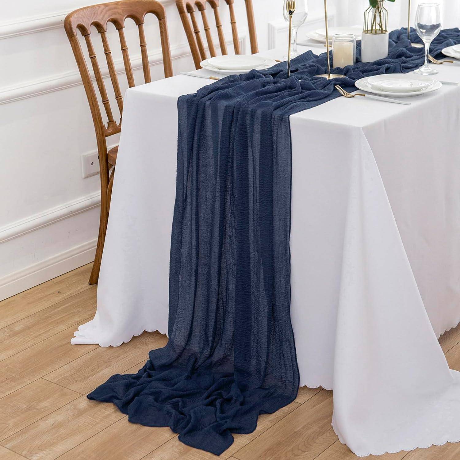 Ein Esstisch, der für eine Mahlzeit gedeckt ist mit Tellern, Gläsern und Besteck. Ein marineblauer Tischläufer drapiert sich elegant über eine weiße Tischdecke.