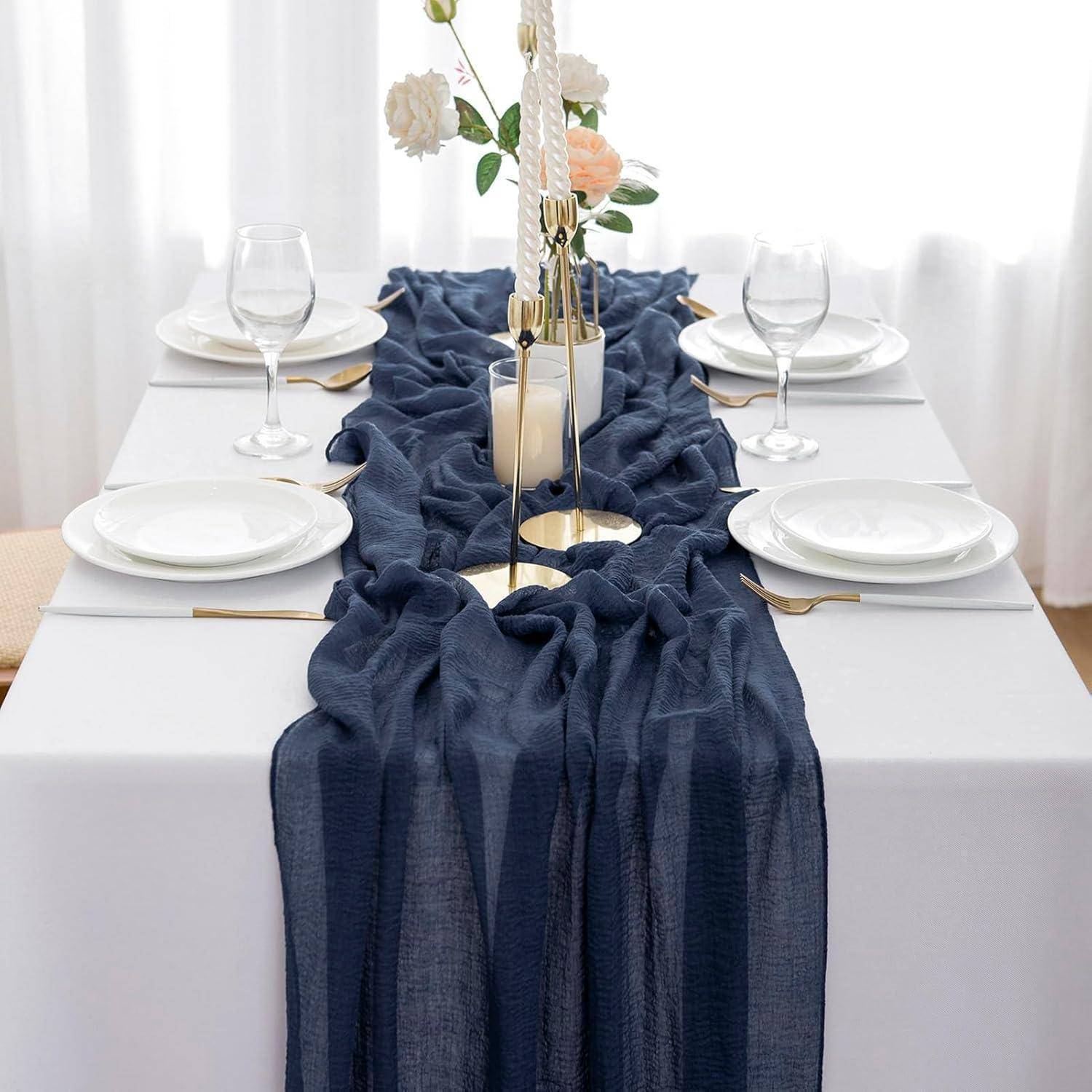 Ein Esstisch, elegant gedeckt mit einer weißen Tischdecke, blauem Läufer, Tellern, Gläsern und einem Blumengesteck mit Kerzen.