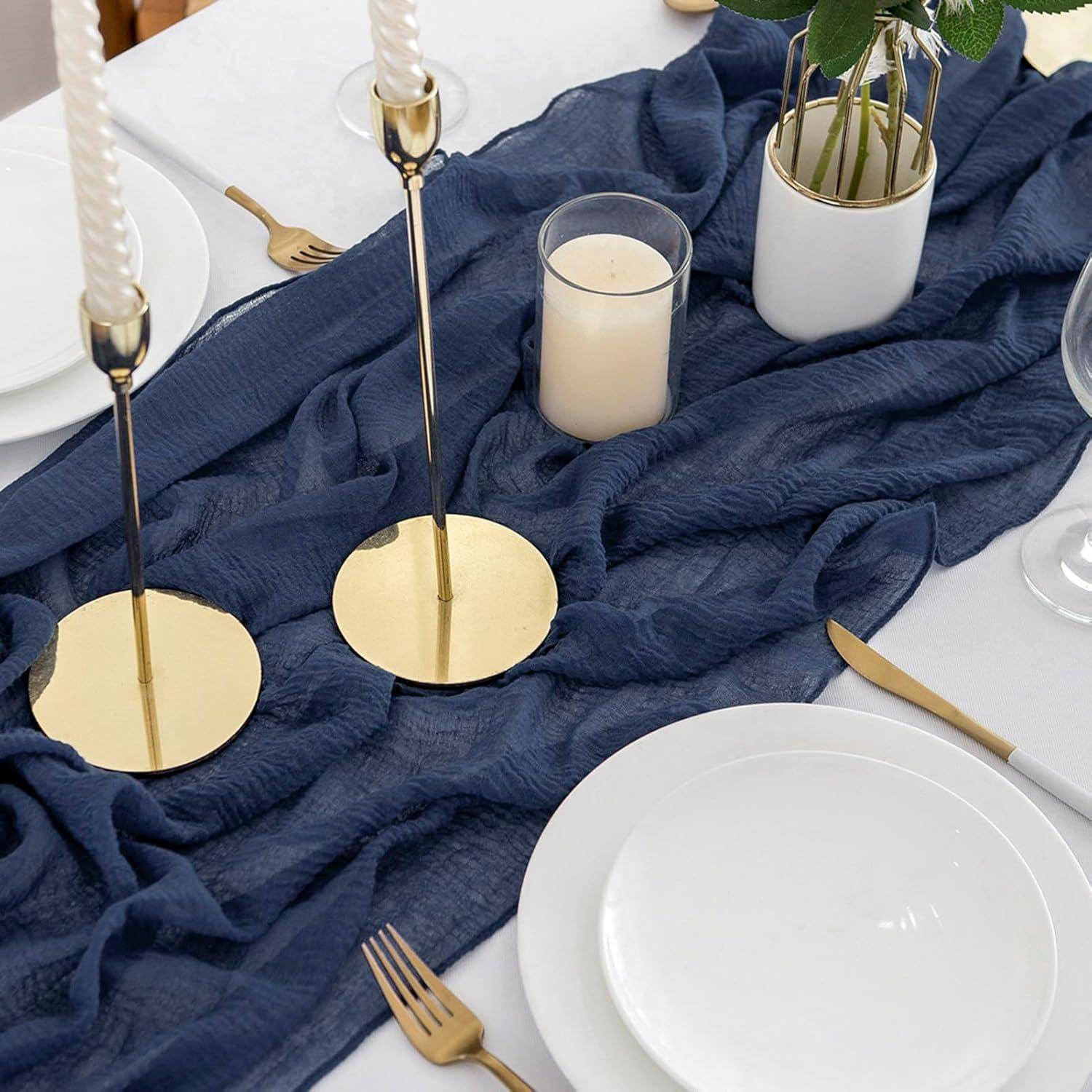 Ein für das Dinner gedeckter Tisch mit blauem Textilläufer, goldenen Kerzenhaltern mit gedrehten Kerzen, einer Topfpflanze und weißen Tellern mit goldenen Bestecken.