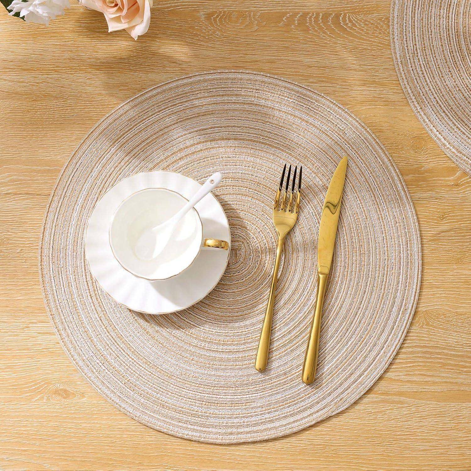Eine weiße Teetasse mit einem Löffel auf einer Untertasse steht auf einem runden geflochtenen Tischset. Eine goldene Gabel und ein goldenes Messer liegen daneben auf einem Holztisch.