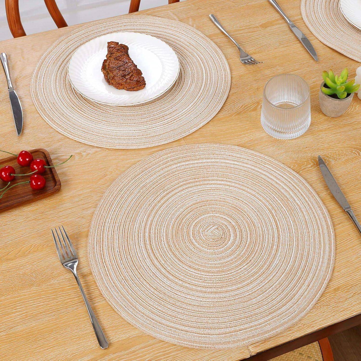 Ein Esstisch für zwei Personen, ausgestattet mit runden beigen Platzdeckchen, einem einzelnen Steak auf einem weißen Teller, Besteck, einem Glas Wasser und einer kleinen Pflanze.