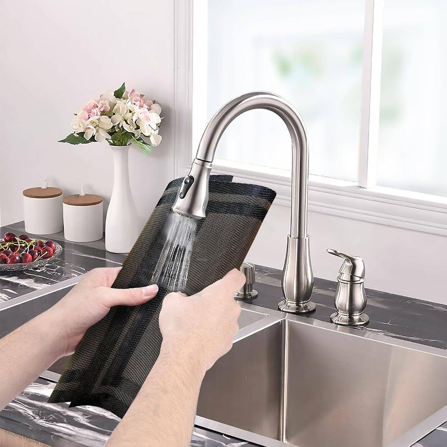 Eine Person spült eine schwarze Küchenmatte unter einem Edelstahl-Wasserhahn in einer modernen Küche mit einer Blumenvase und Behältern im Hintergrund.