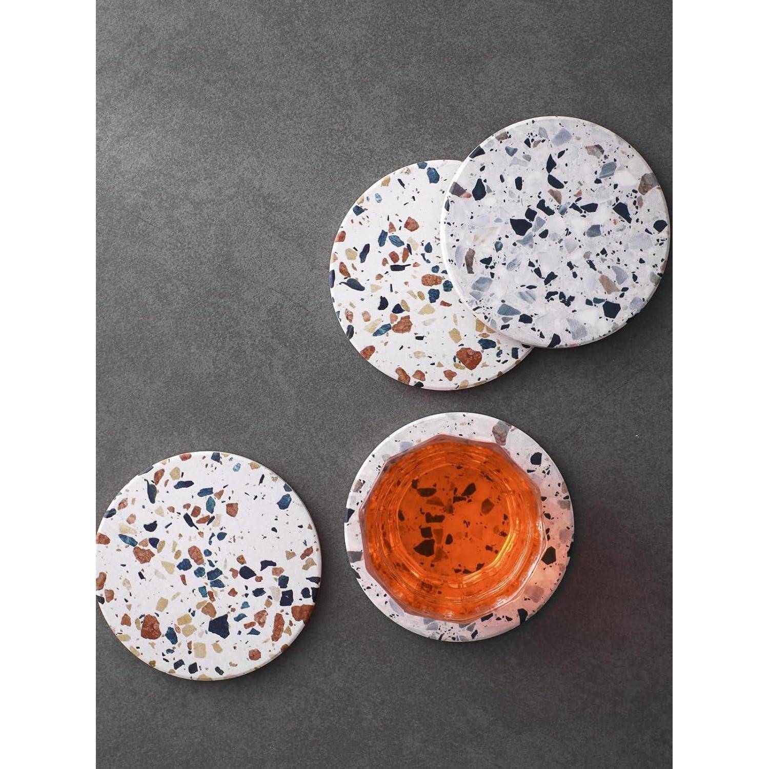 Drei Terrazzo-Untersetzer in verschiedenen Positionen, einer davon mit einem Glas mit bernsteinfarbener Flüssigkeit, auf einer dunkelgrauen Oberfläche liegend.