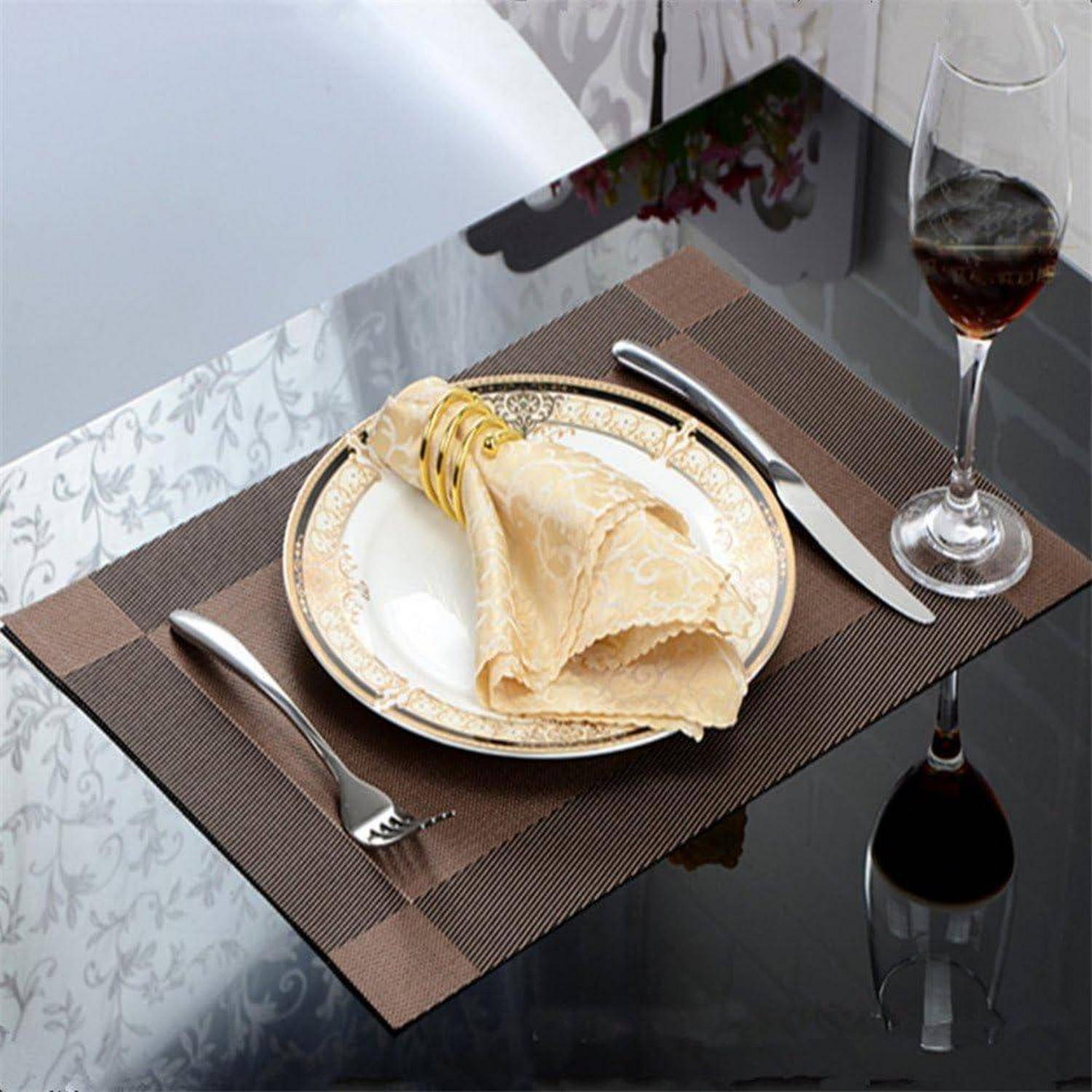 Elegante Tischgedeck mit einem gemusterten Teller, in einem Ring gefalteter beiger Serviette, Besteck und einem Weinglas auf einer reflektierenden Oberfläche.