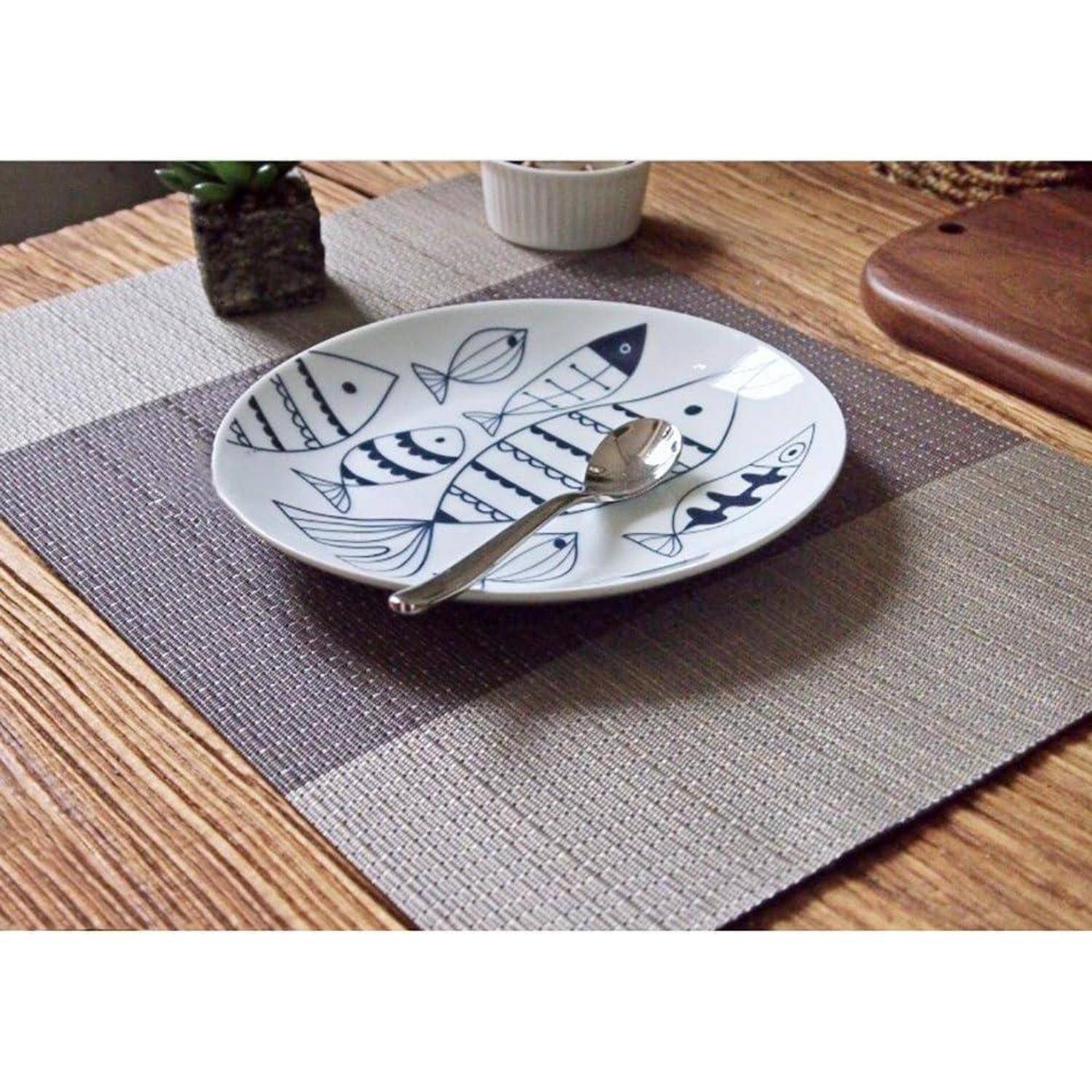 Ein gemusterter Teller mit blauen Fischdesigns steht auf einem geflochtenen Platzset und hält einen Löffel. Der Hintergrund umfasst eine kleine Pflanze und ein Gefäß.